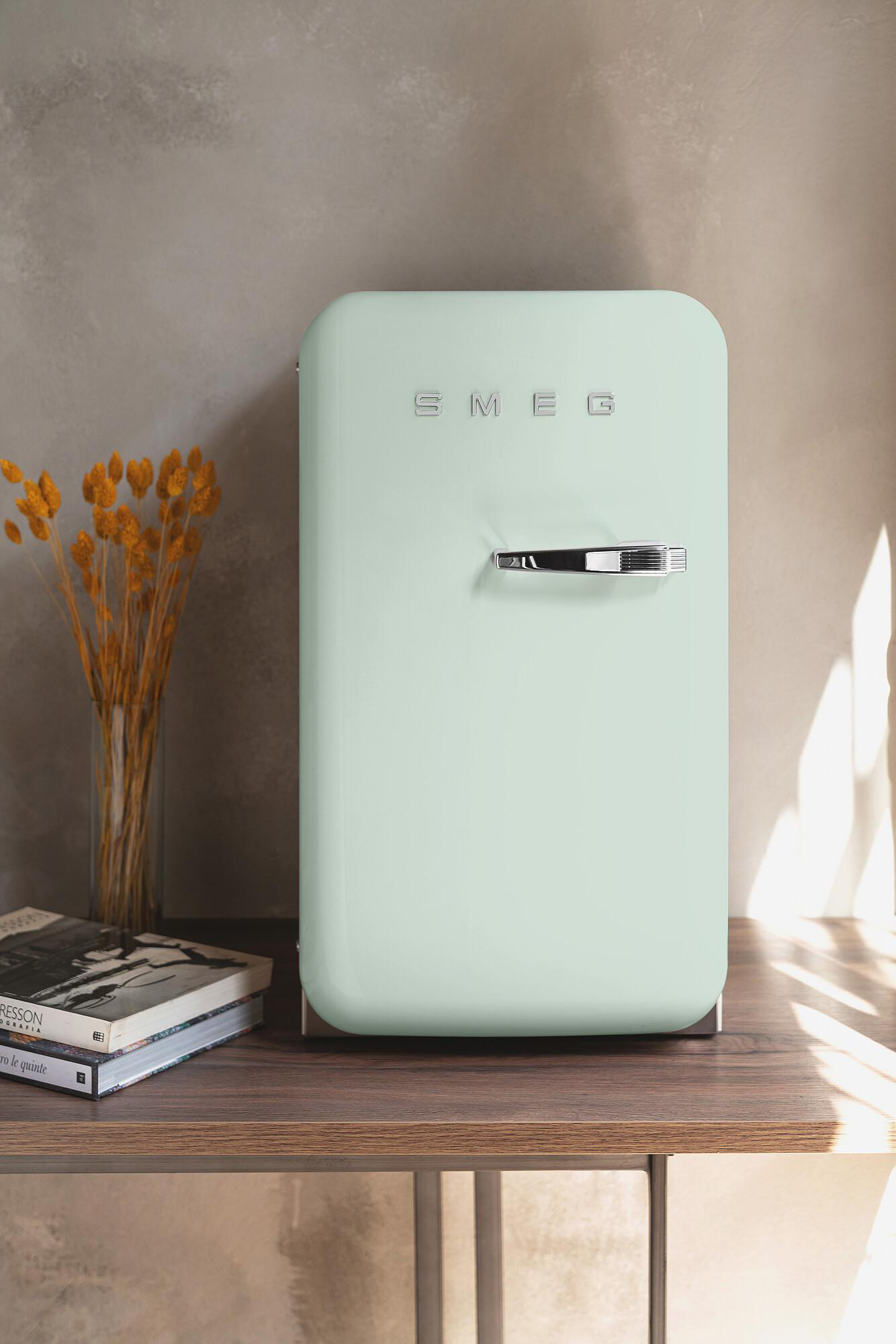 Ein mintgrüner Smeg-Kühlschrank auf einem Holztisch. Daneben eine Vase mit gelben Trockenblumen und Büchern. Der Hintergrund ist grau.