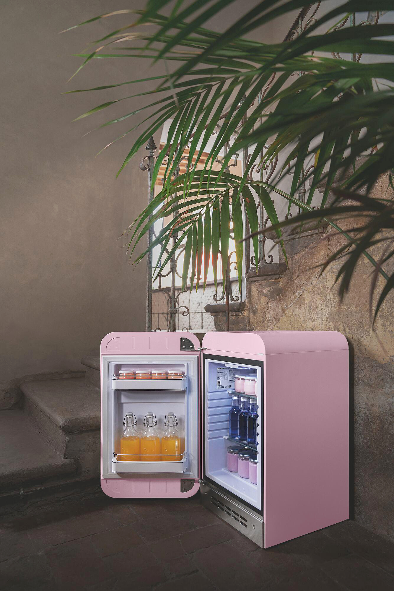 Pinkfarbener Retro-Kühlschrank, geöffnet, mit Getränken. Palmfarn oben und Steinstufen links. Schmiedeeisernes Tor und Wand im Hintergrund.