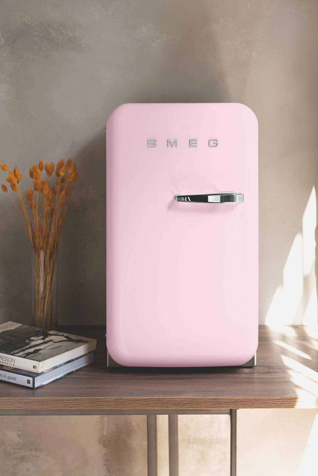 Ein rosafarbener Smeg-Minikühlschrank steht auf einem Holzregal. Bücher und eine Vase mit trockenen Blumen stehen auf dem Regal. Der Hintergrund ist eine Wand.