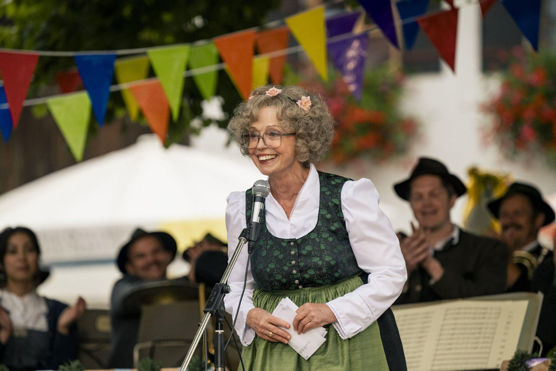 Eine Frau in Tracht spricht in ein Mikrofon und lächelt. Dahinter spielt eine Band, und bunte Fahnen hängen darüber.
