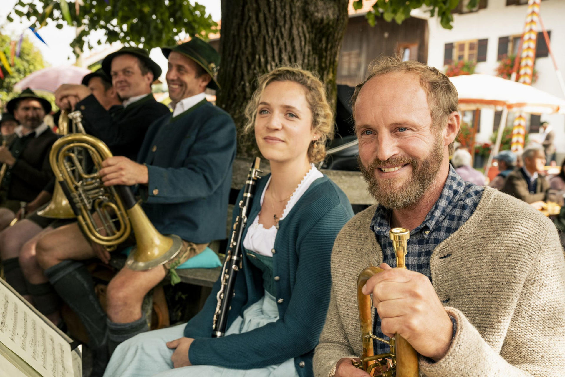 Eine Blaskapelle spielt. Eine Frau spielt Klarinette. Leute tragen Tracht und lächeln. Eine sonnige Szene im Freien.