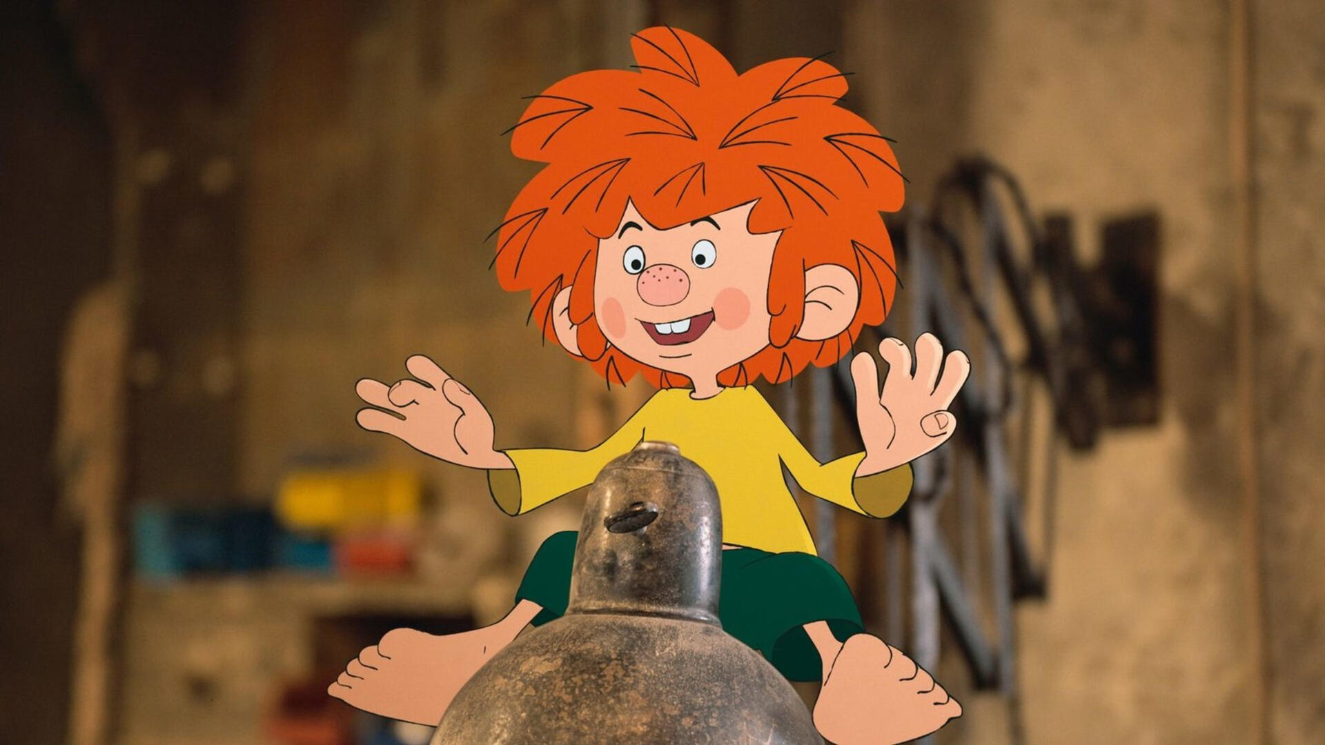 Ein Cartoon-Junge mit roten Haaren, gelbem Hemd und grünen Shorts sitzt mit offenen Armen auf einem Metallobjekt. Der Hintergrund ist unscharf.