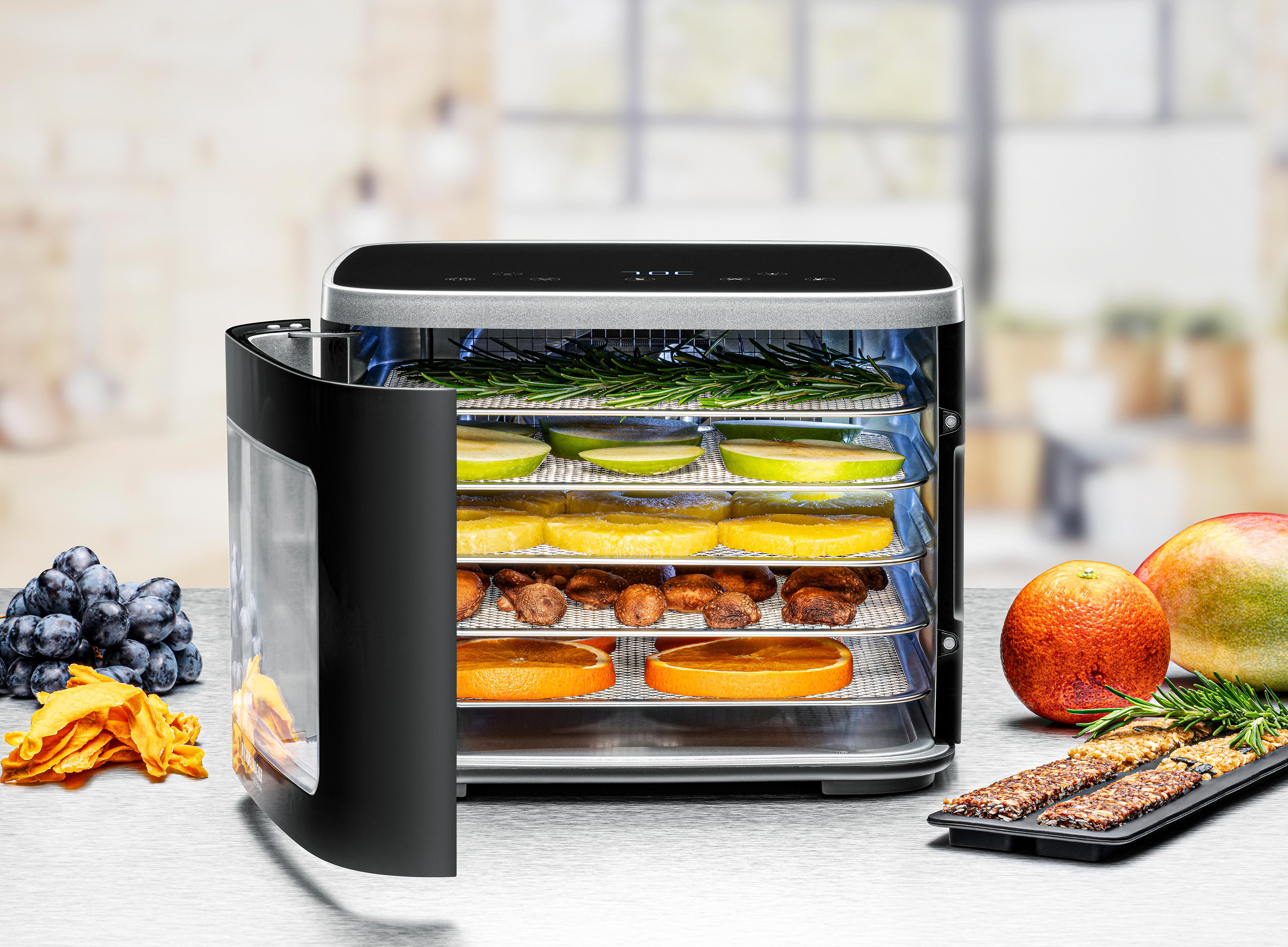 Lebensmitteltrockner mit verschiedenen Früchten und Kräutern, wie Äpfeln, Orangen und Mangos. Trauben und getrocknete Mangoscheiben daneben.