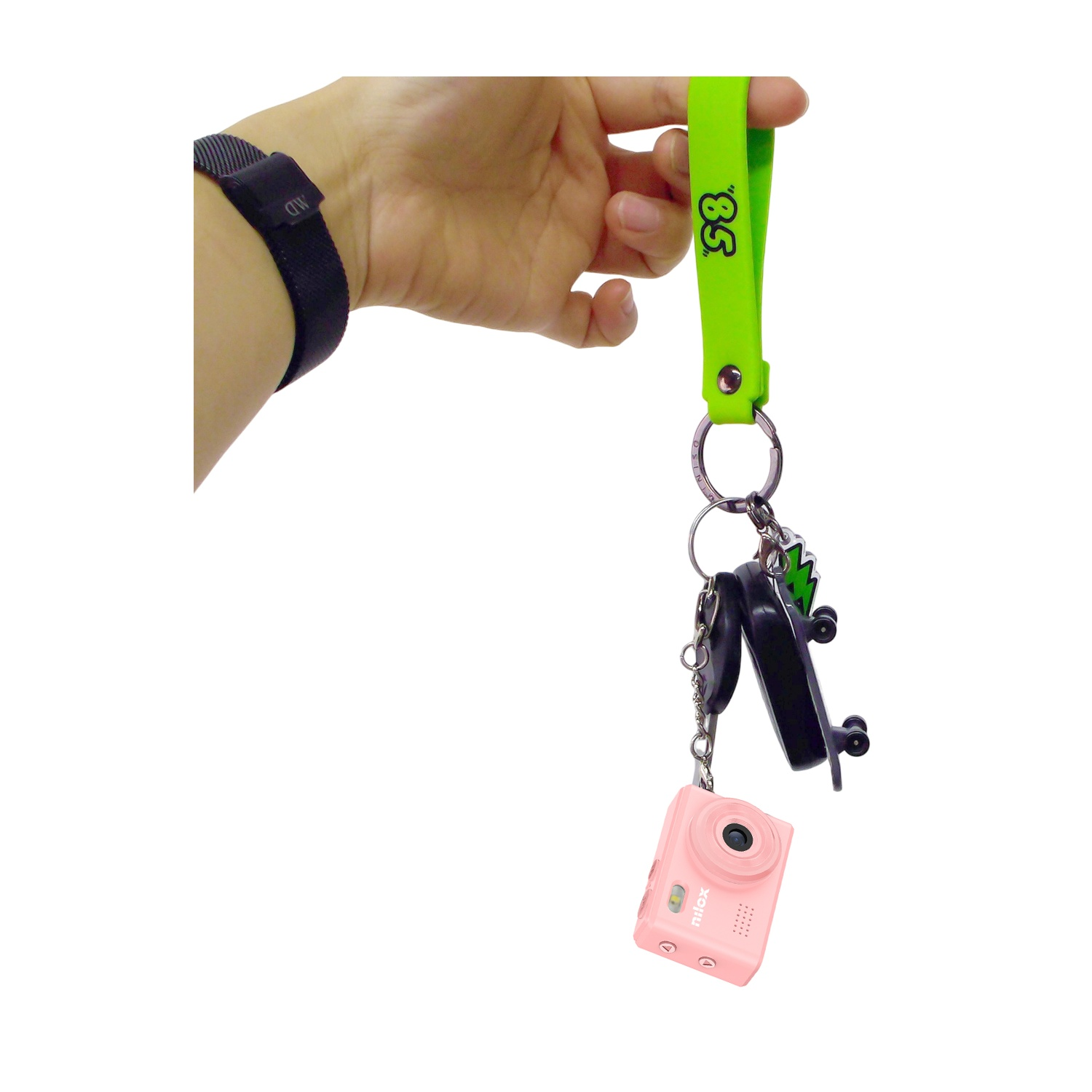 Mano che tiene un cordino verde neon con chiavi, una fotocamera rosa e un ciondolo a forma di skateboard nero.
