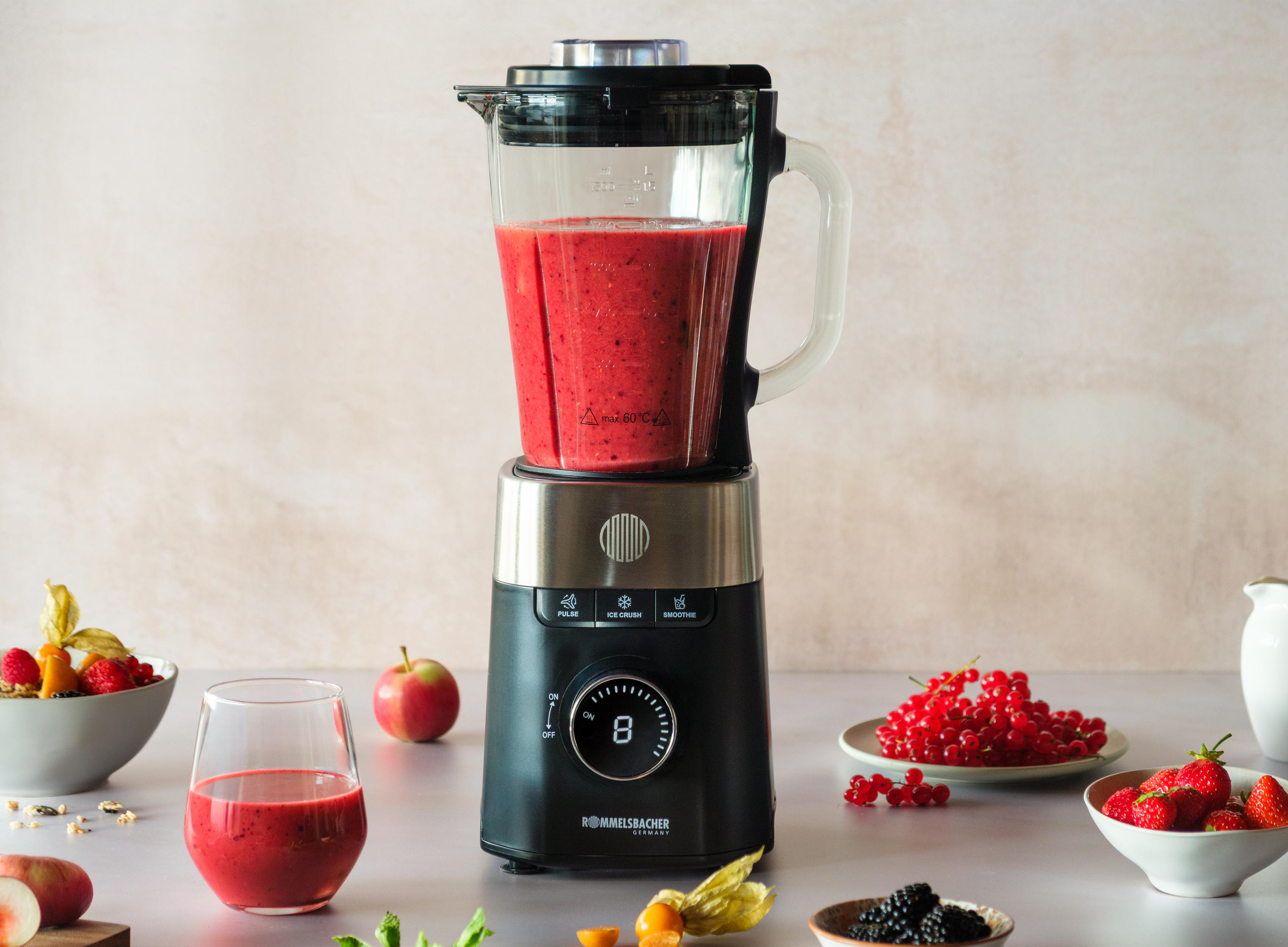 Ein Mixer mit rotem Smoothie, Früchten wie Erdbeeren und Johannisbeeren auf einem Tisch mit einem Glas Smoothie.