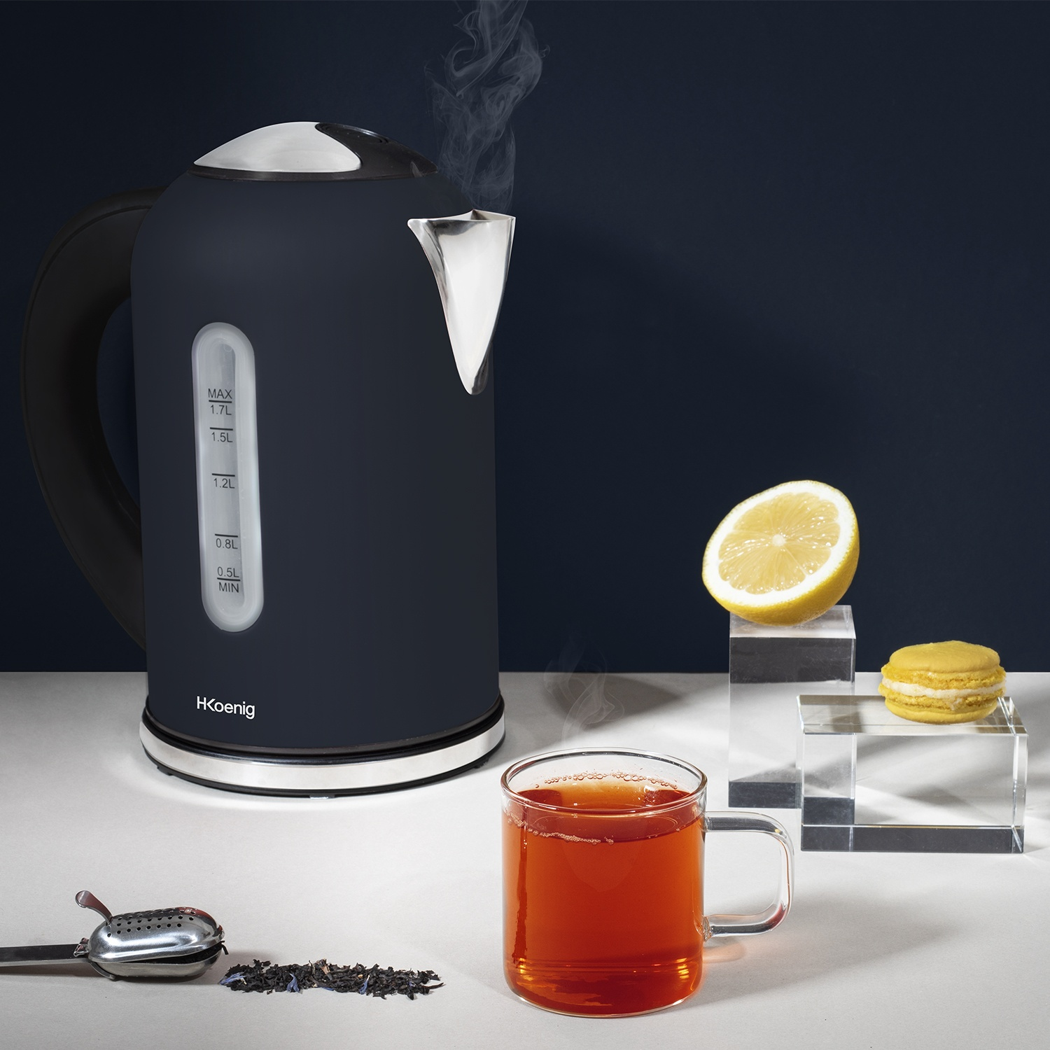 Un bollitore nero emette vapore accanto a una tazza di tè, una metà di limone e un macaron. Foglie di tè e un infusore di tè in primo piano.