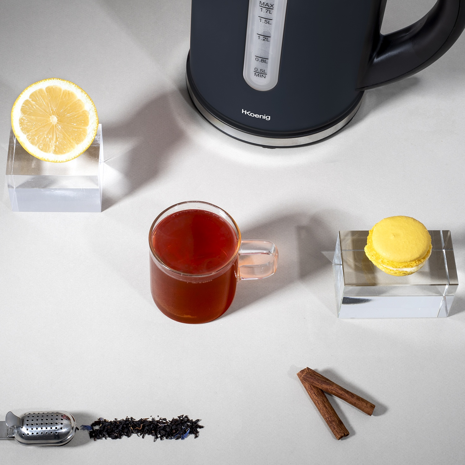 Una natura morta con tazza di tè, fetta di limone, macaron, bollitore, foglie di tè e bastoncini di cannella su superficie bianca.