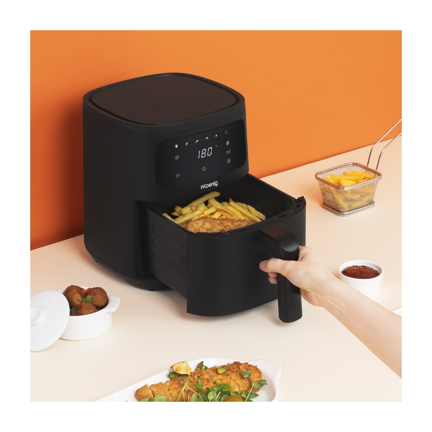 Friggitrice ad aria nera con patatine fritte e cibo. Una mano tiene il cestello. Altri cibi sono sul tavolo con salsa di pomodoro.