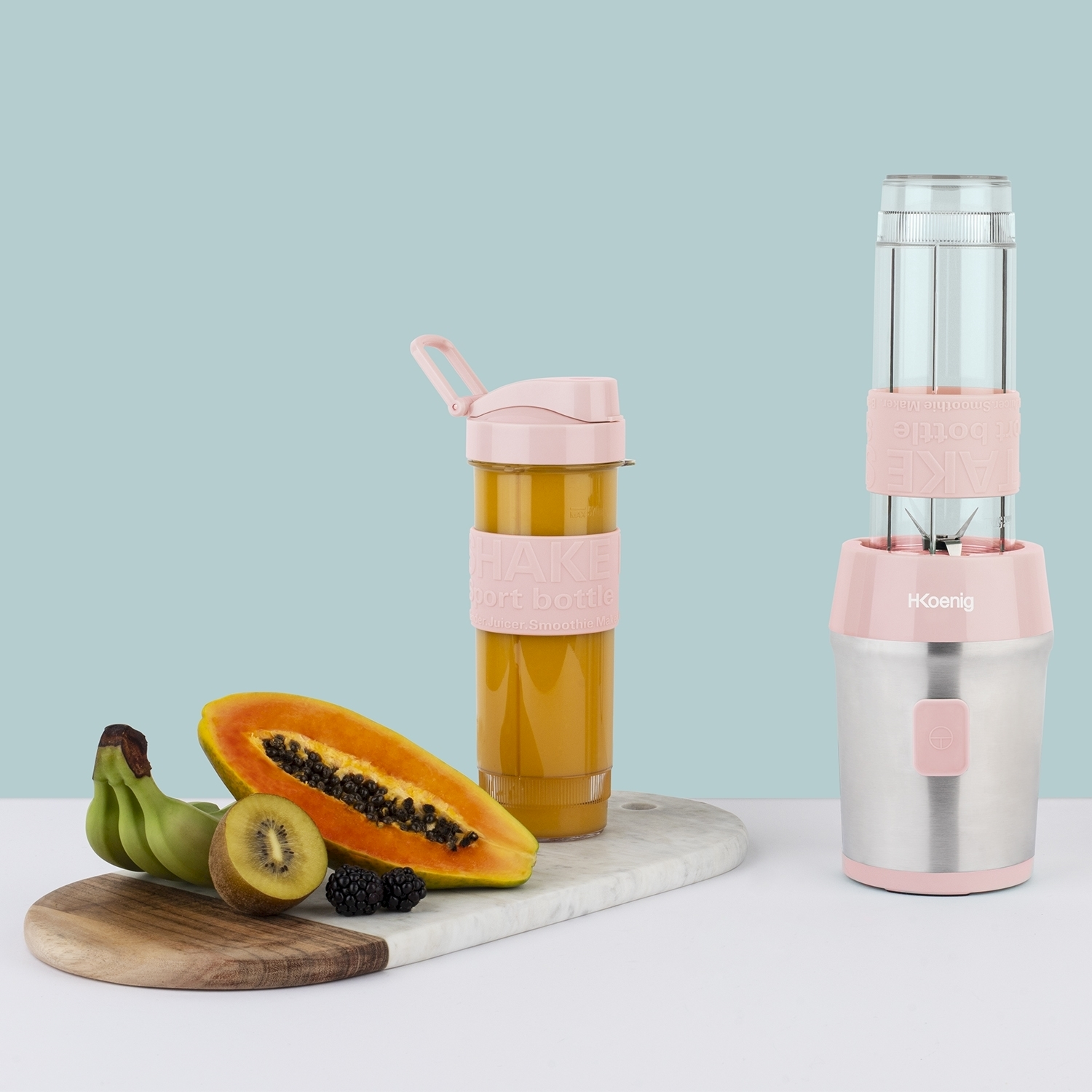Un frullatore e una bottiglia shaker con frutta come banana, papaya e kiwi su una superficie bianca, sfondo blu.
