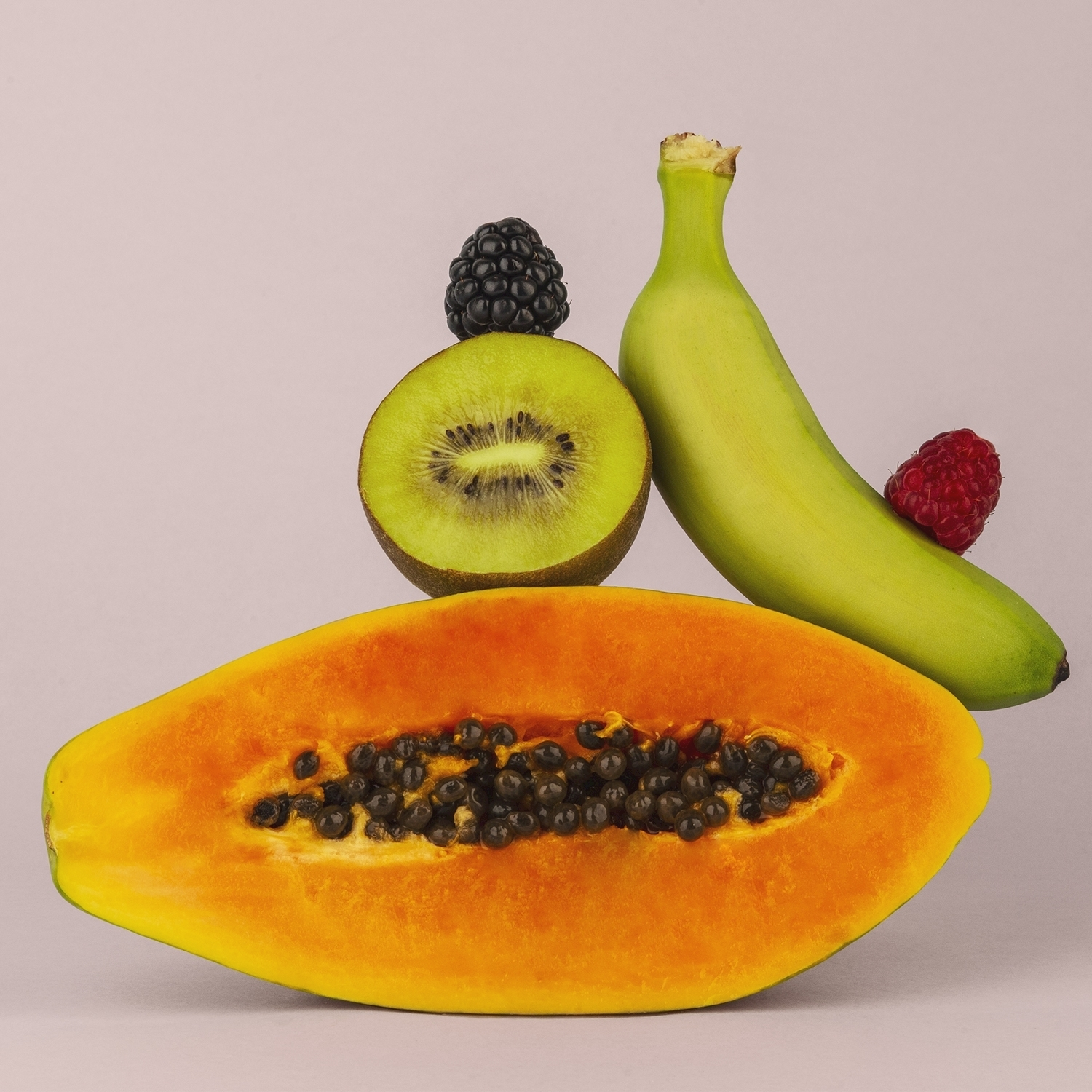 Una papaya, un kiwi, una banana, un lampone e una mora sono disposti su una superficie lilla chiaro. La frutta è fresca e colorata.