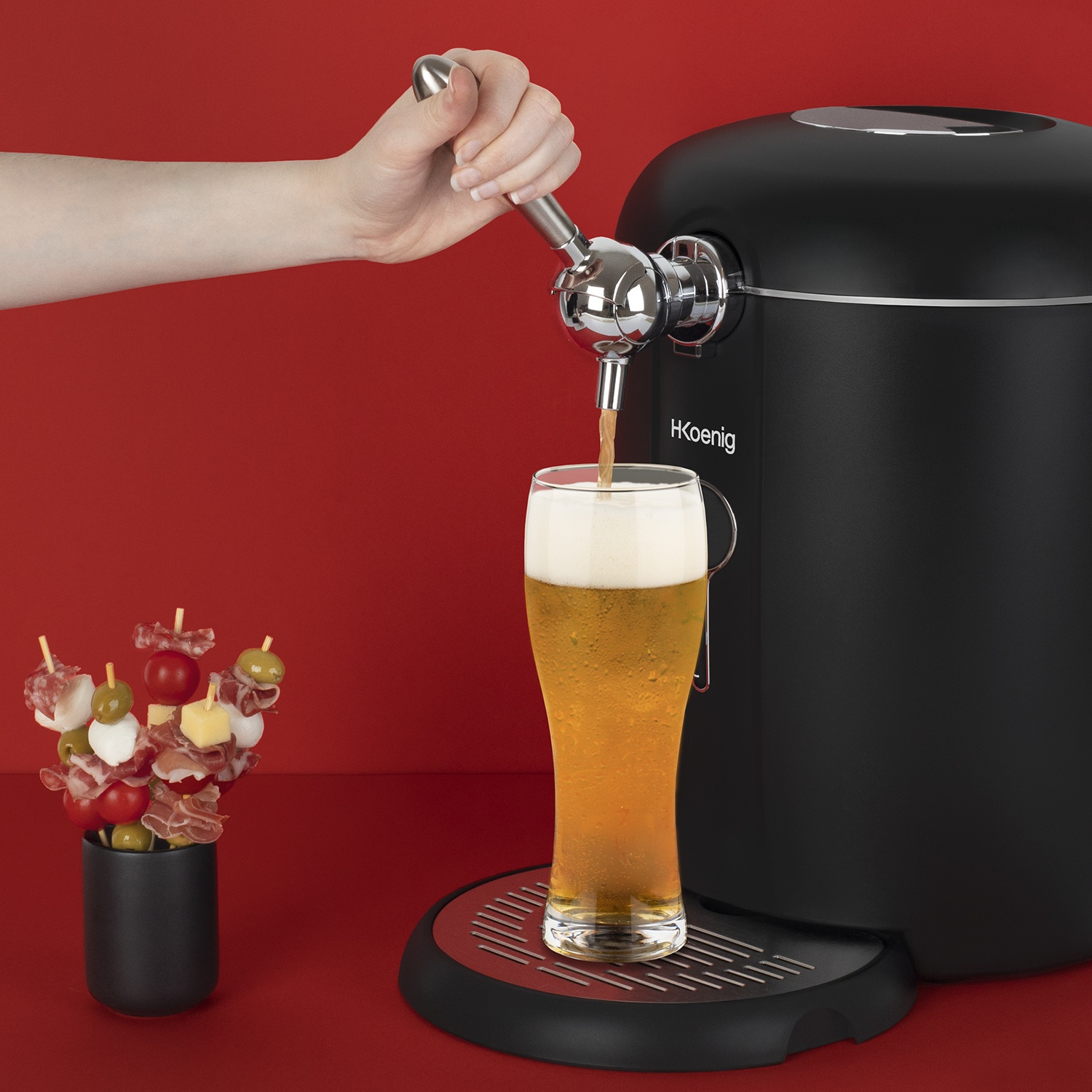 Una mano versa birra da un rubinetto in un bicchiere. Un distributore di birra nero è accanto al bicchiere e spiedini con cibo a sinistra.
