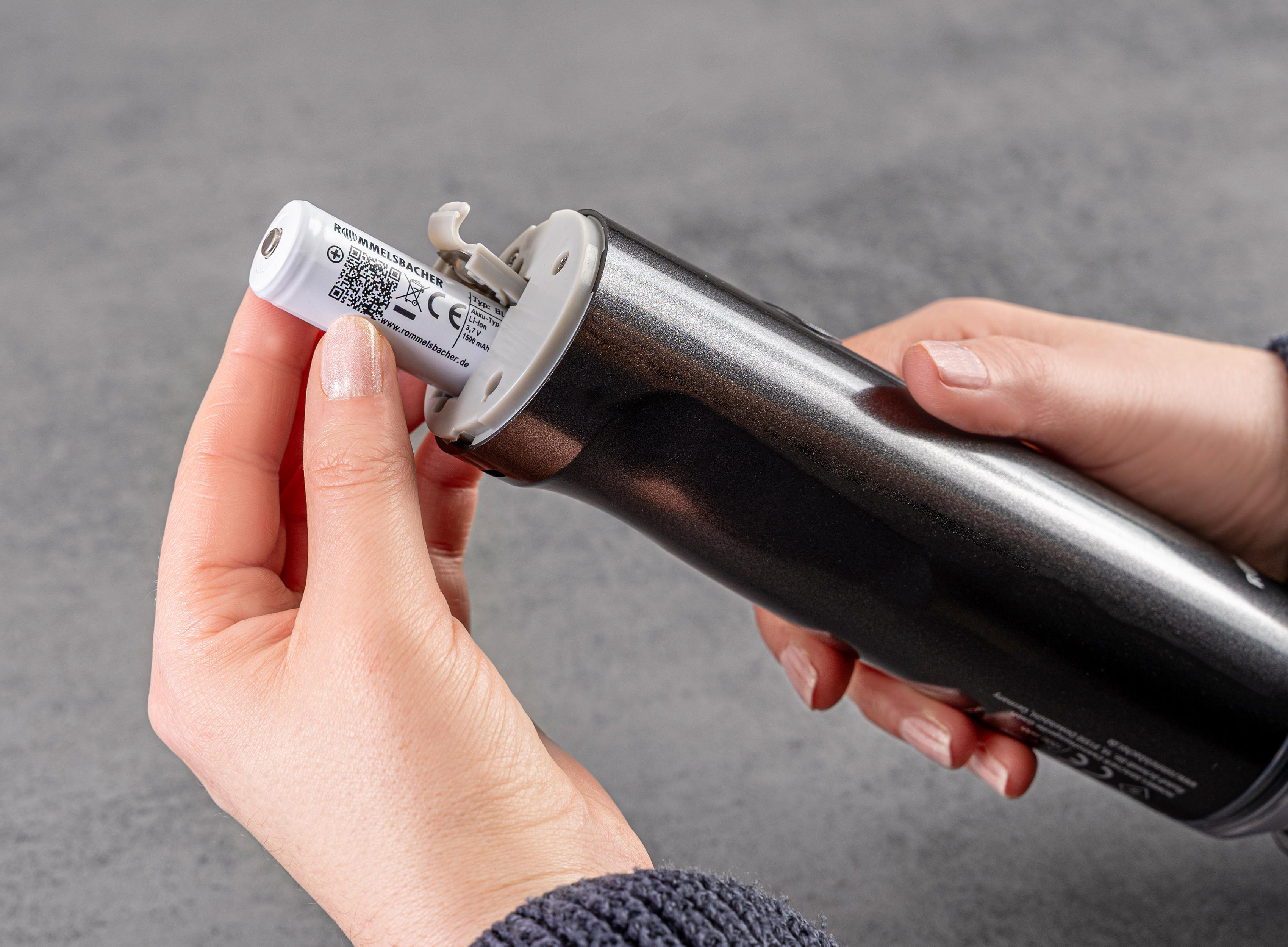 Hände legen eine Batterie in einen dunkelgrauen Handmixer ein. Die Batterie ist weiß mit schwarzem Text.