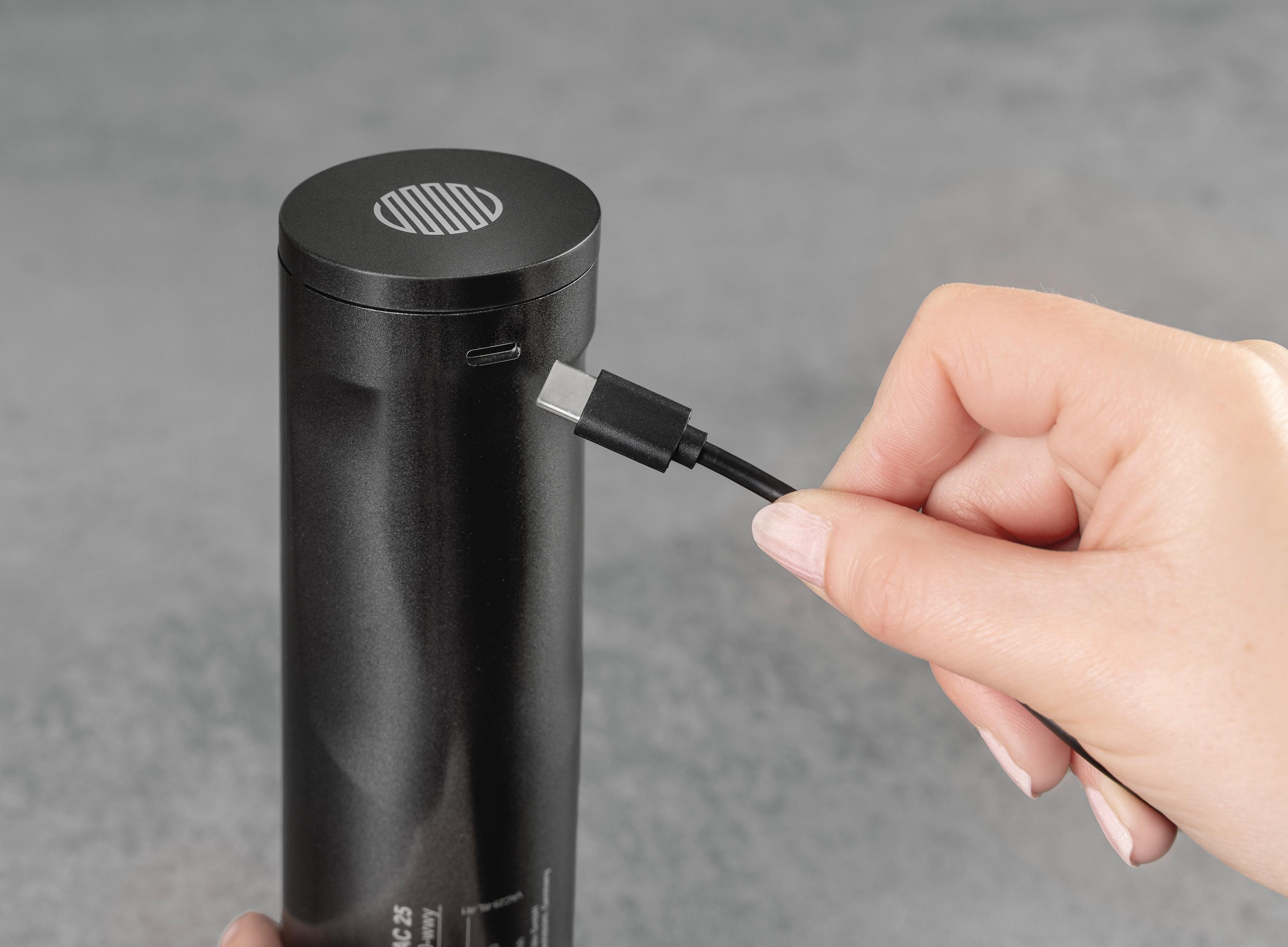 Eine Hand hält ein schwarzes USB-C-Kabel und verbindet es mit einem schwarzen Zylinder. Der Hintergrund ist grau.