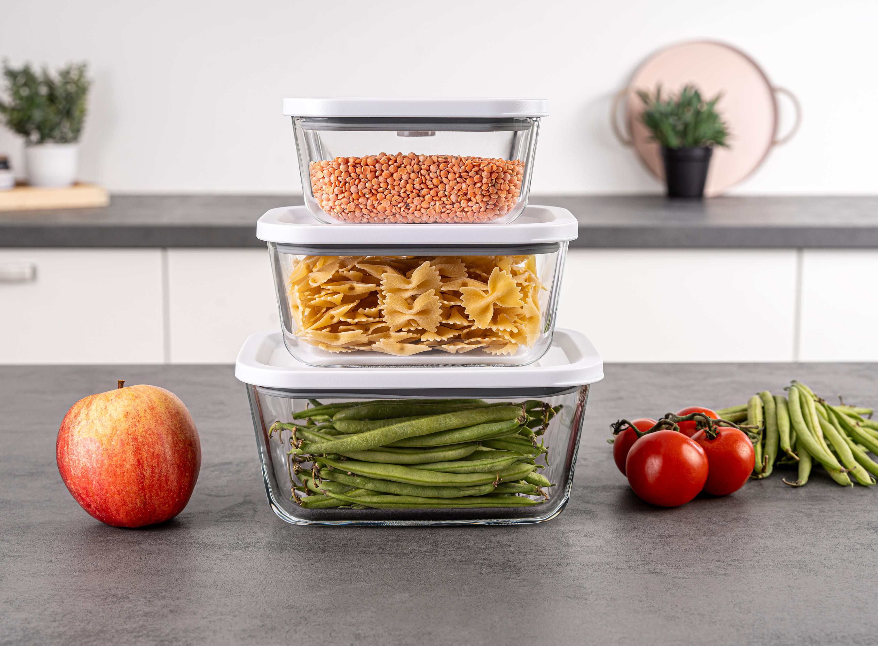 Drei Glasbehälter mit Linsen, Nudeln und grünen Bohnen. Ein Apfel und Tomaten sind ebenfalls vorhanden.