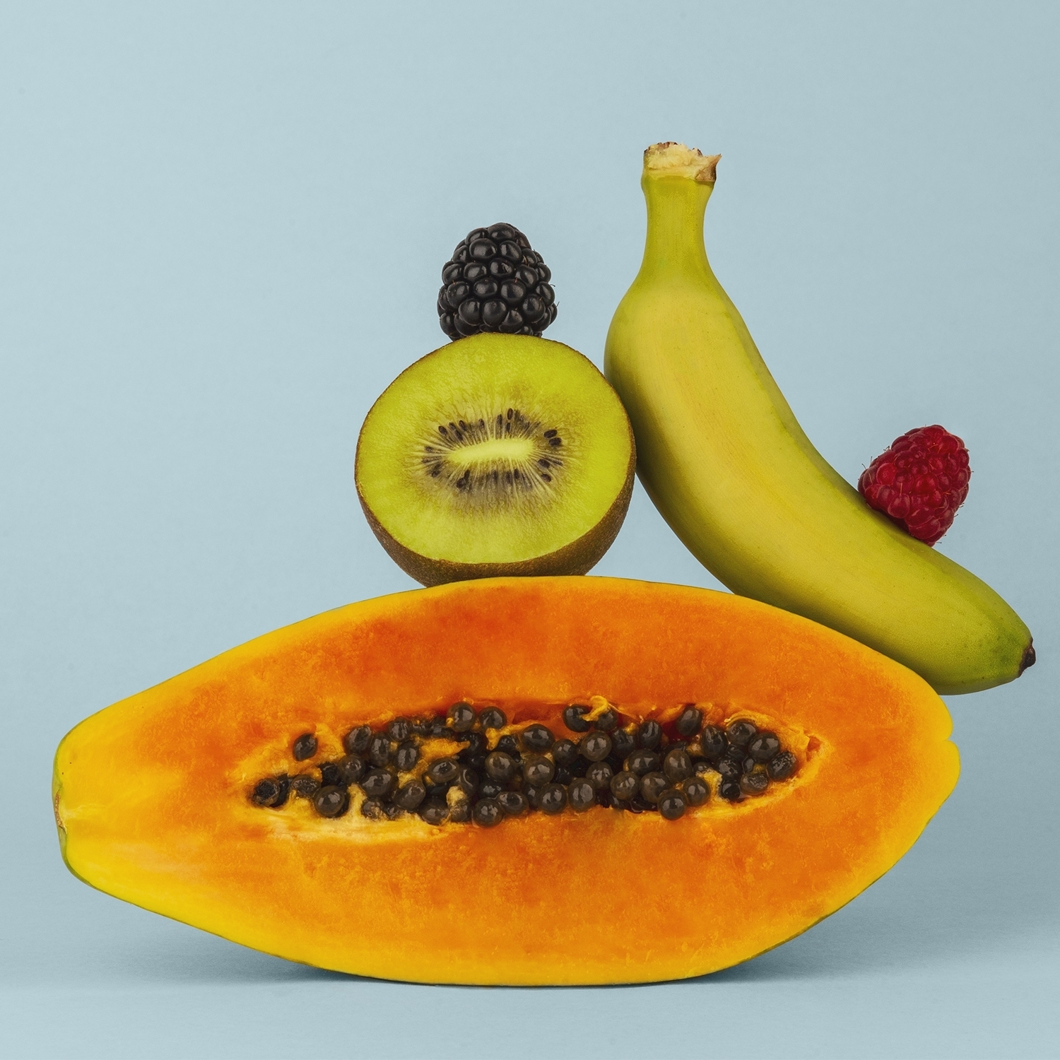 Metà papaya, kiwi, banana, mora e lampone su sfondo azzurro. La frutta è disposta artisticamente.