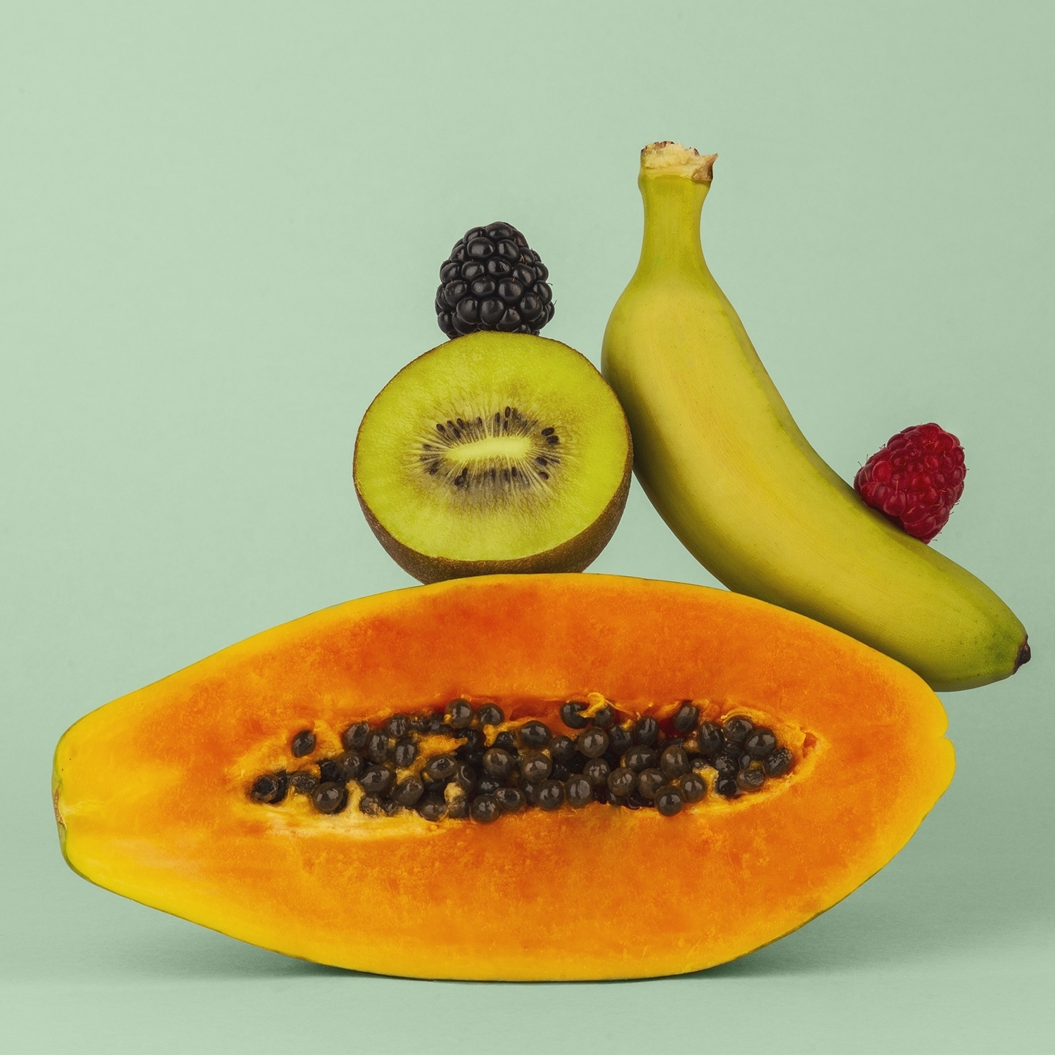 Metà papaya, kiwi, banana, mora e lampone su sfondo verde chiaro. Frutta disposta in una composizione equilibrata.