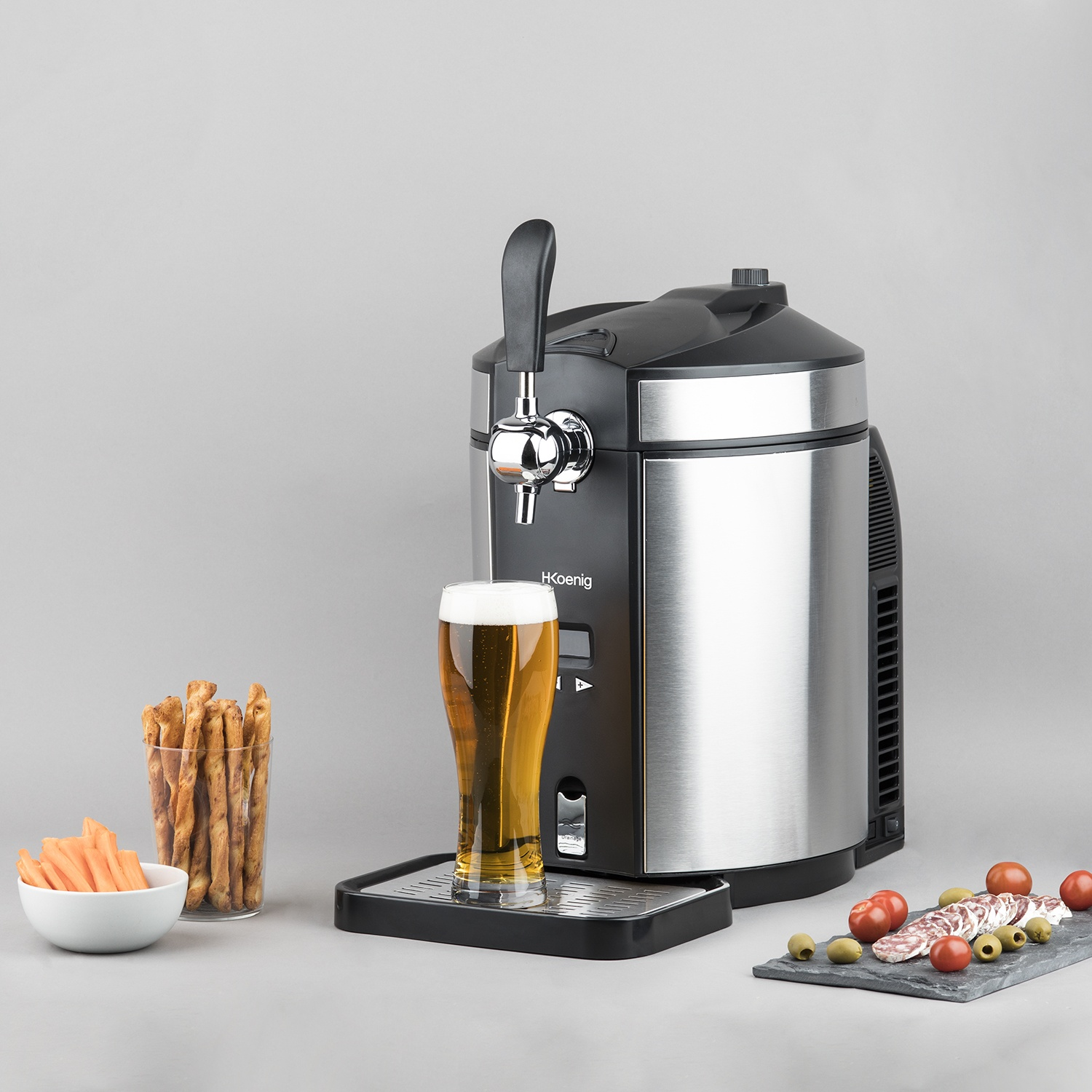 Un rubinetto della birra con un bicchiere di birra, grissini, bastoncini di carote, salame, olive e pomodori su sfondo grigio.