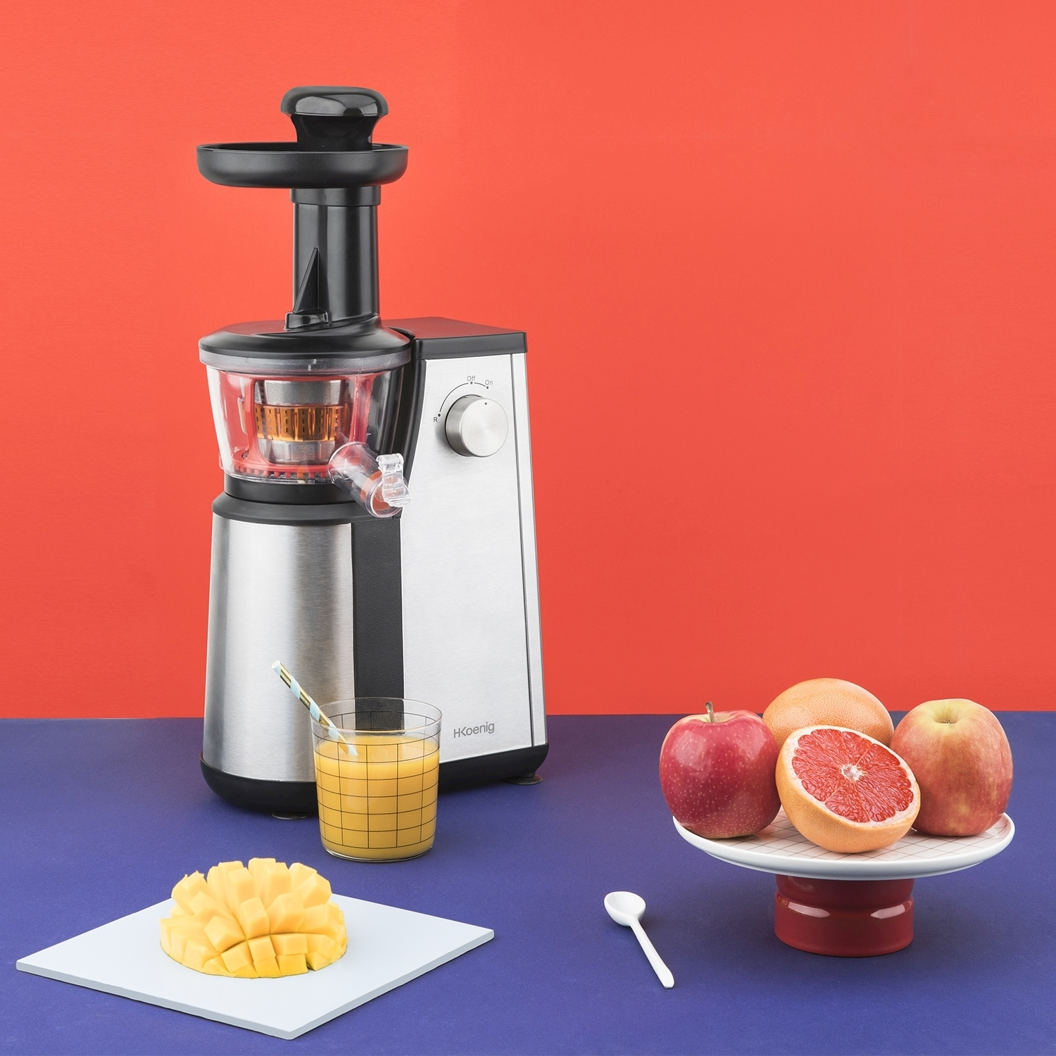 Centrifuga, bicchiere di succo, mango e frutta su un supporto. Sfondo rosso e blu.