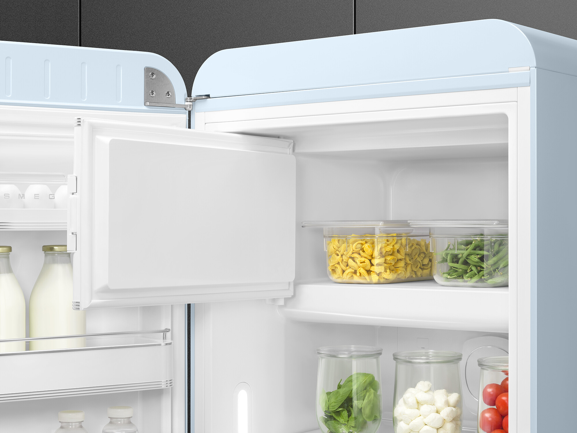 Offener Kühlschrank gefüllt mit Lebensmitteln: Pasta, grüne Bohnen, Basilikum, Tomaten, Mozzarella und Milch. Hellblaue Tür.