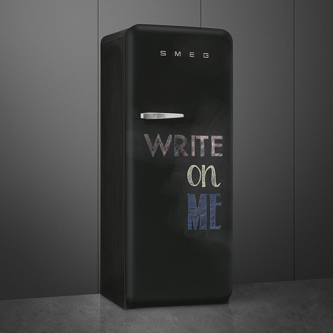 Schwarzer SMEG-Kühlschrank mit der Aufschrift 'WRITE ON ME'. Er steht vor einer dunkelgrauen Wand und auf einem grauen Boden.