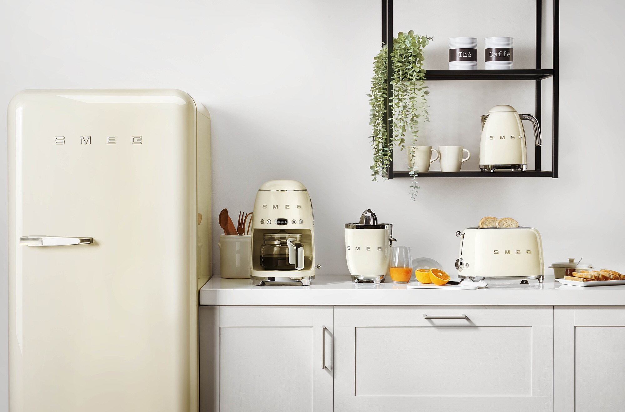 Cremefarbene Küchengeräte auf einer Theke: Kühlschrank, Kaffeemaschine, Entsafter, Toaster, Wasserkocher. Orangensaft, Brot und Tassen sind vorhanden.