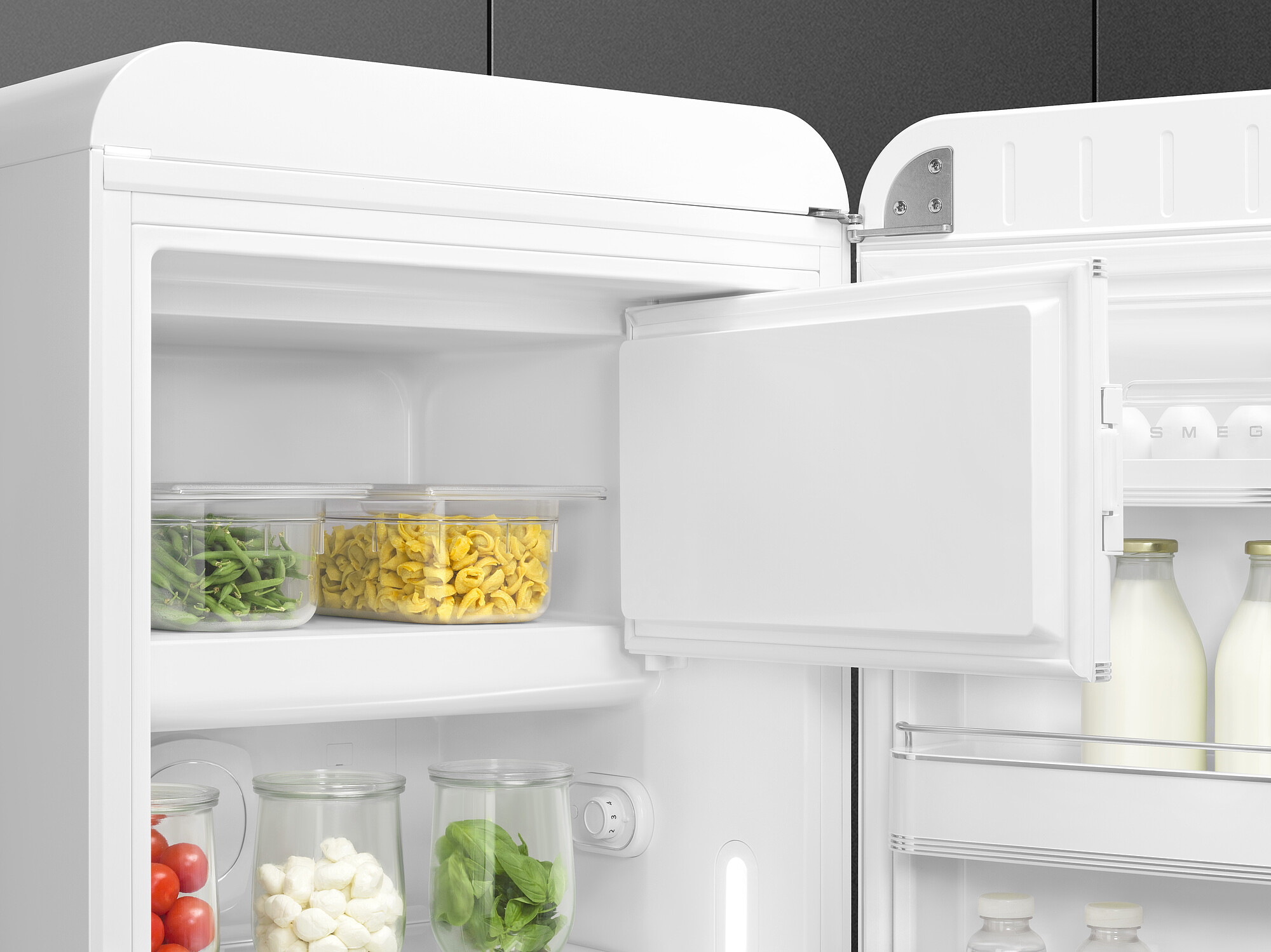 Offener weißer Kühlschrank mit Lebensmitteln gefüllt. Enthält grüne Bohnen, Tortellini, Tomaten, Mozzarella, Basilikum und Milchflaschen.