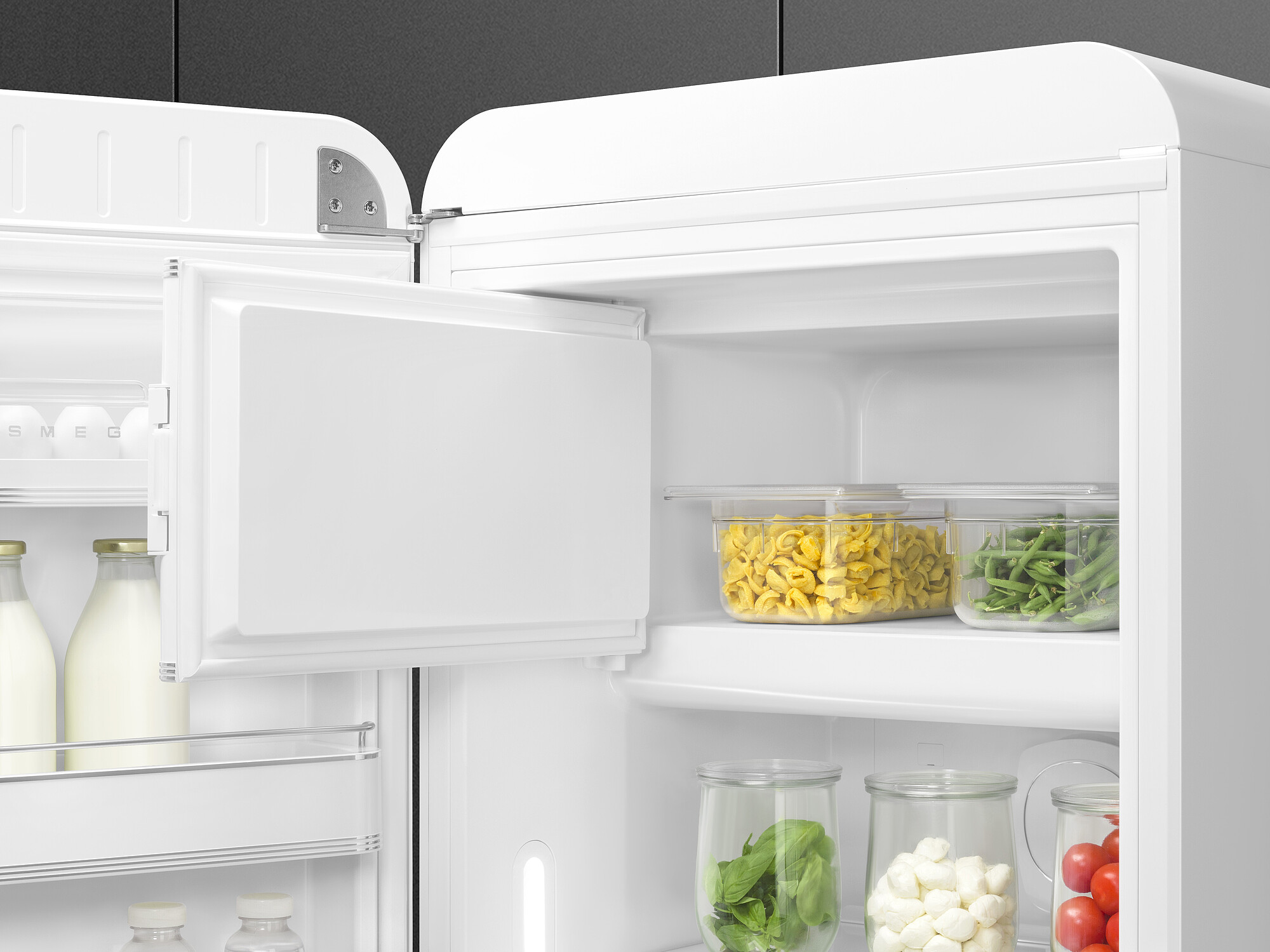 Offener weißer Kühlschrank mit Lebensmitteln: Nudeln, grüne Bohnen, Basilikum, Mozzarella, Tomaten und Milchflaschen. Eierablage an der Tür.