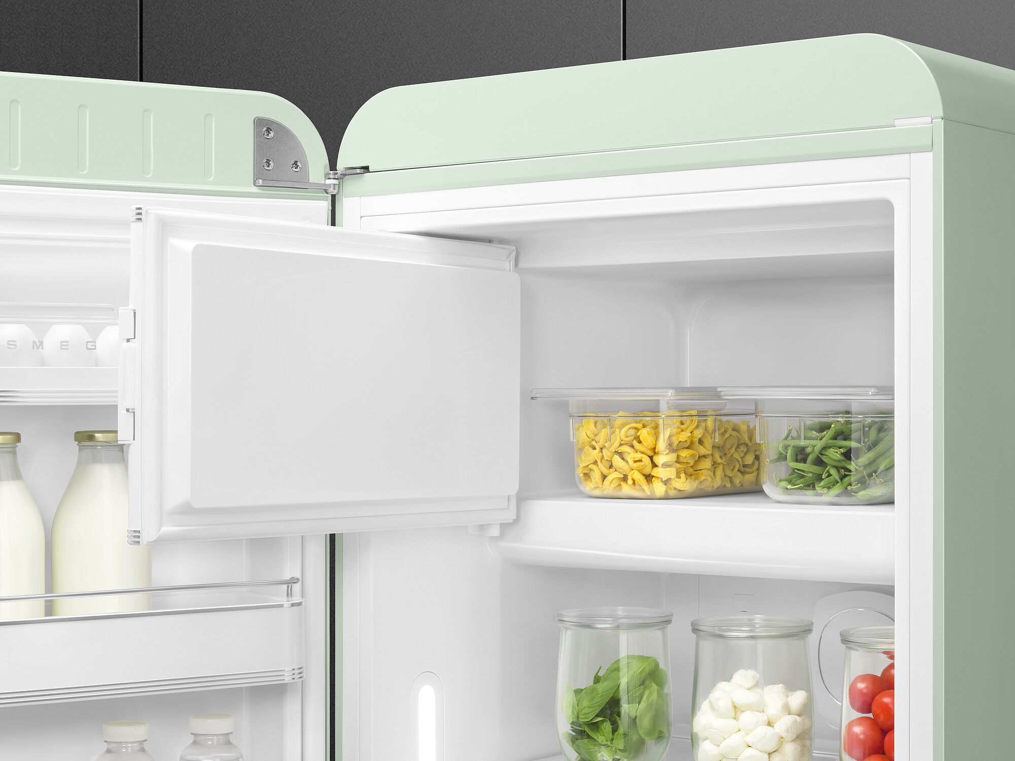 Offener mintgrüner Kühlschrank. Im Inneren befinden sich Lebensmittel: Nudeln, Bohnen, Tomaten, Basilikum und Mozzarella. Milch und Eier sind in der Tür.