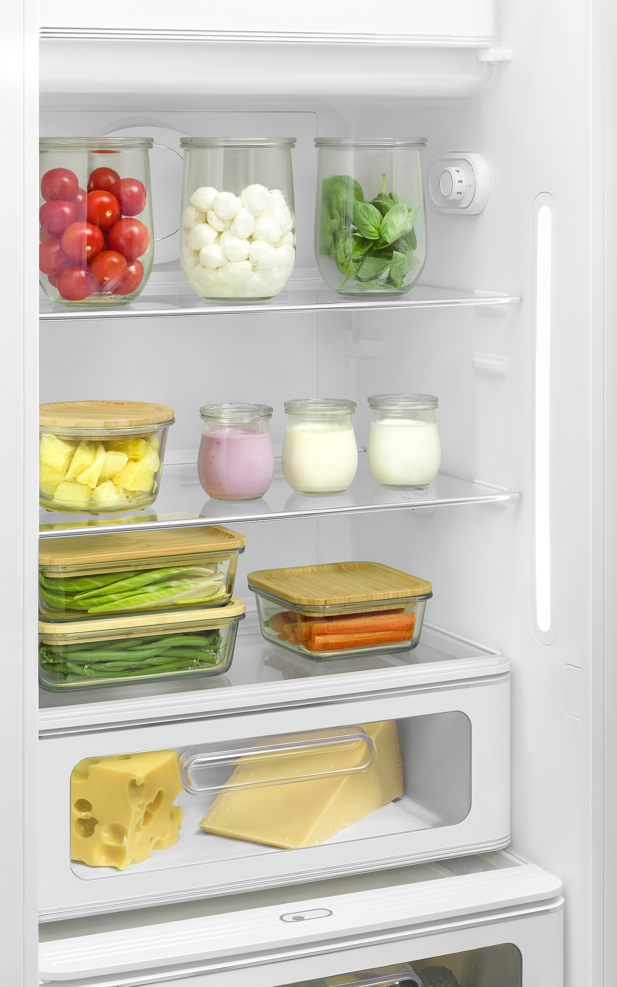 Im Kühlschrank: Tomaten, Mozzarella, Basilikum, Ananas, Joghurt, Karotten, Käse und grüne Bohnen, gelagert in Glasbehältern.