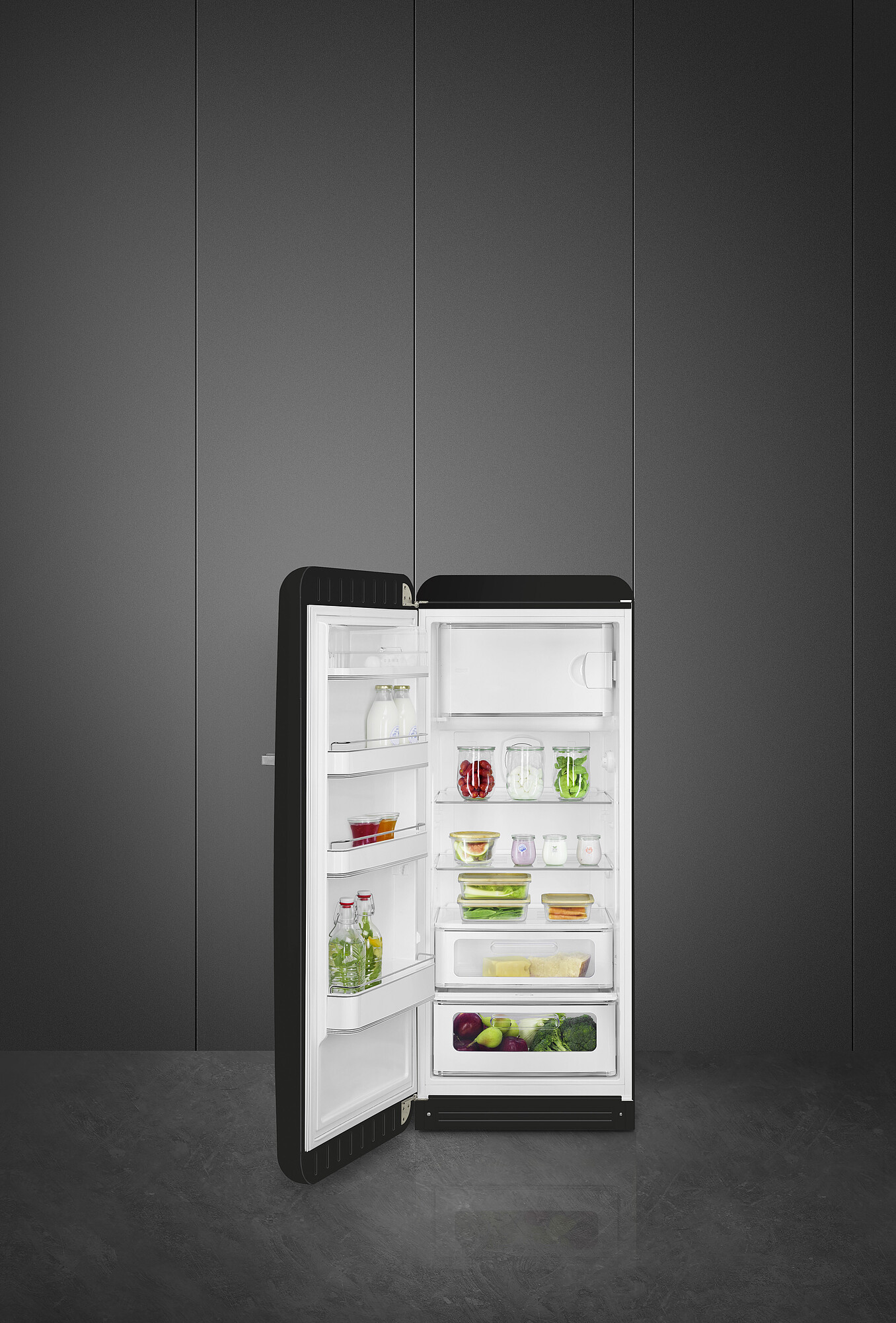 Offener schwarzer Kühlschrank gefüllt mit Obst, Gemüse und Gläsern. Vor einer grauen Wand und Boden.