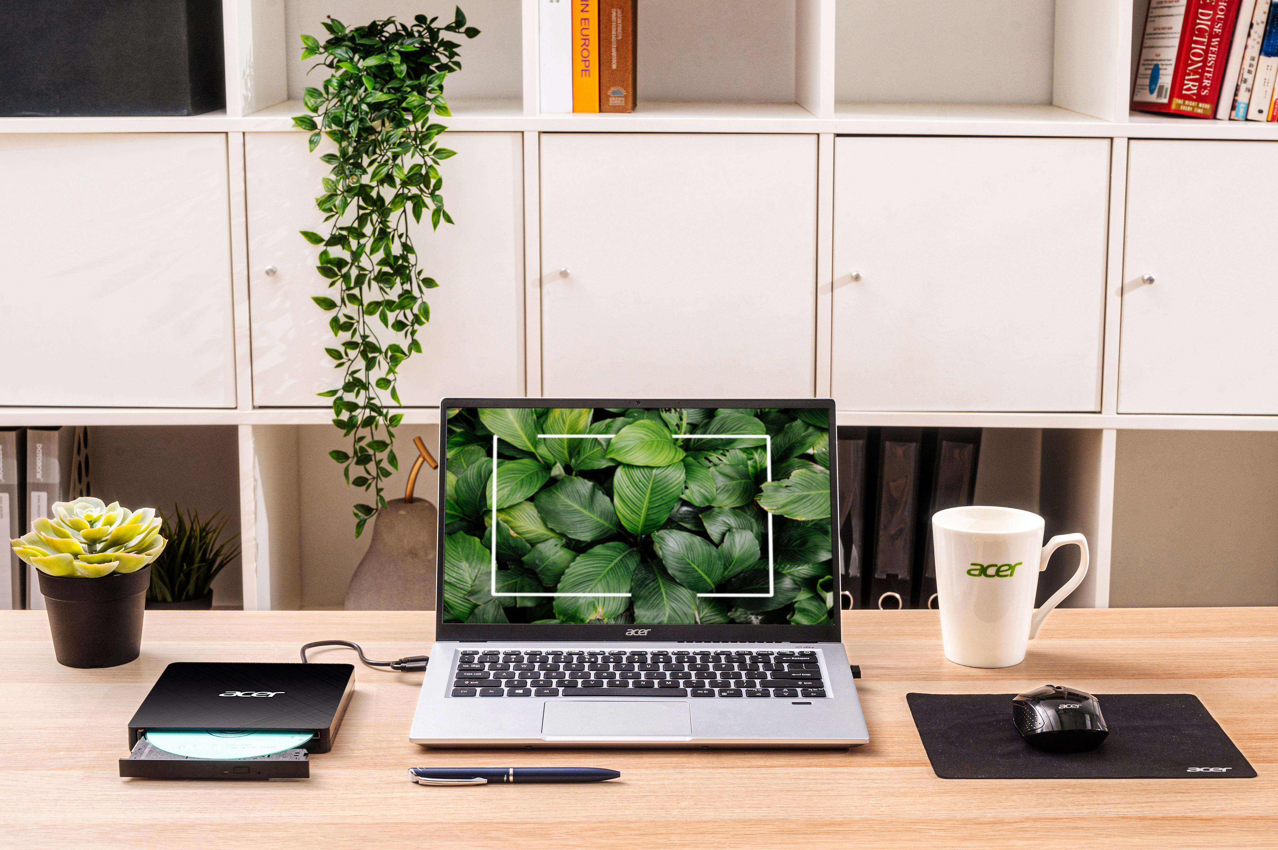 Ein Schreibtisch mit einem Acer-Laptop, einer Tasse, einer Maus und einer Pflanze. Der Laptop zeigt Blätter. Links ein schwarzes CD-Laufwerk.