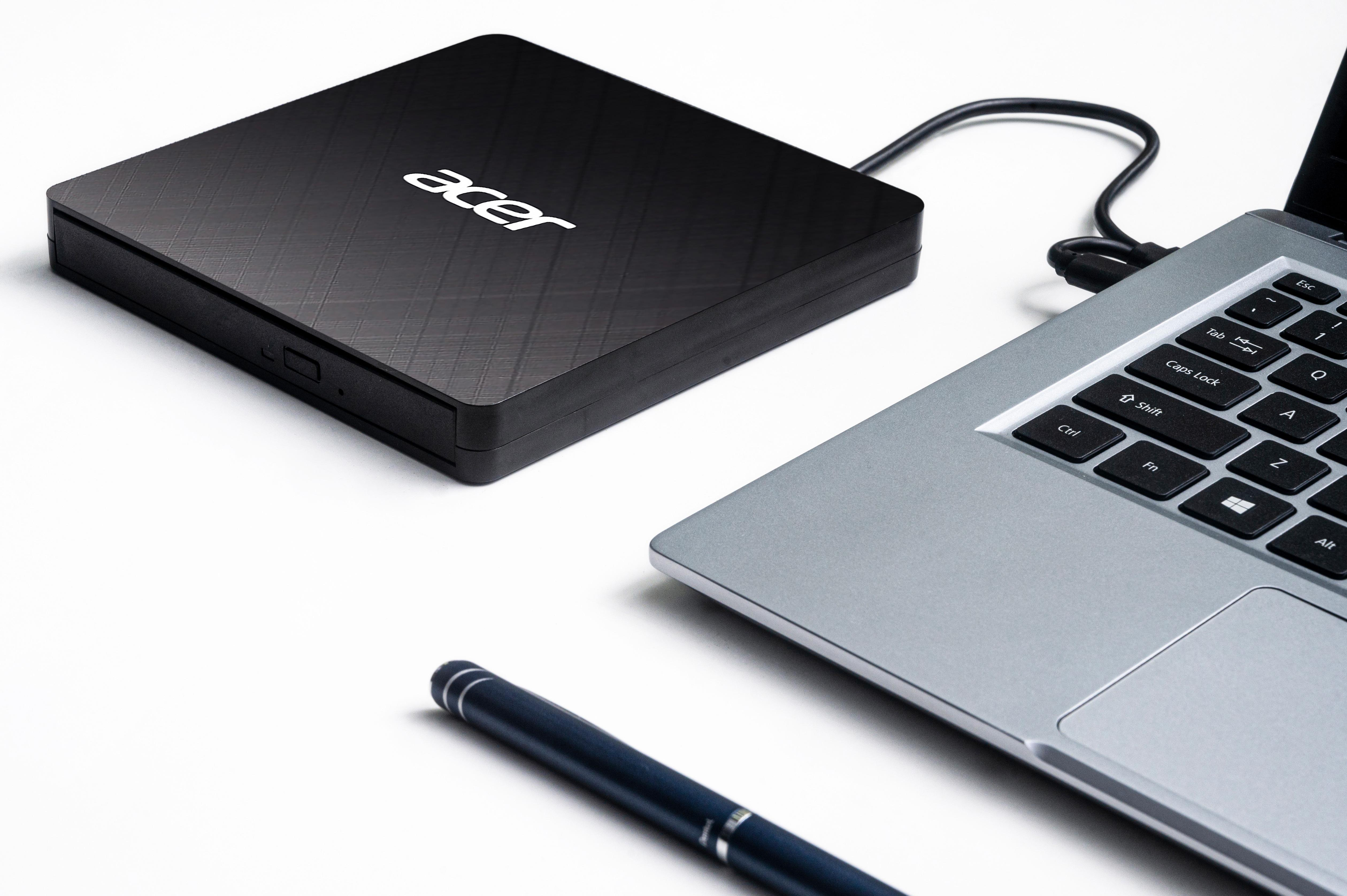 Ein schwarzes Acer externes DVD-Laufwerk neben einem Laptop. Ein Stift befindet sich im Vordergrund. Alles auf weißem Hintergrund.