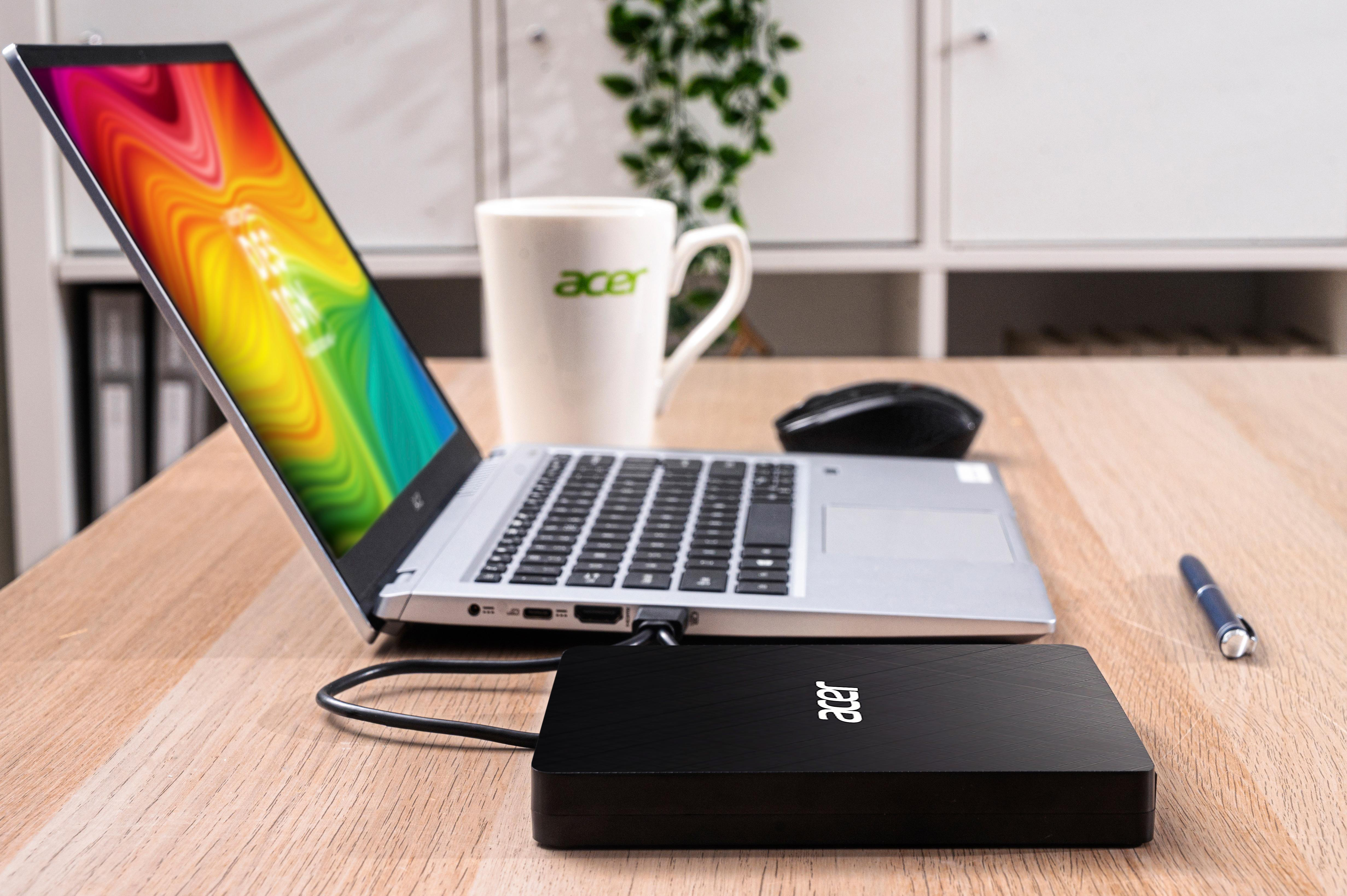Laptop, externe Festplatte, Becher und Stift auf einem Holztisch. Der Laptop-Bildschirm zeigt ein farbenfrohes Bild.