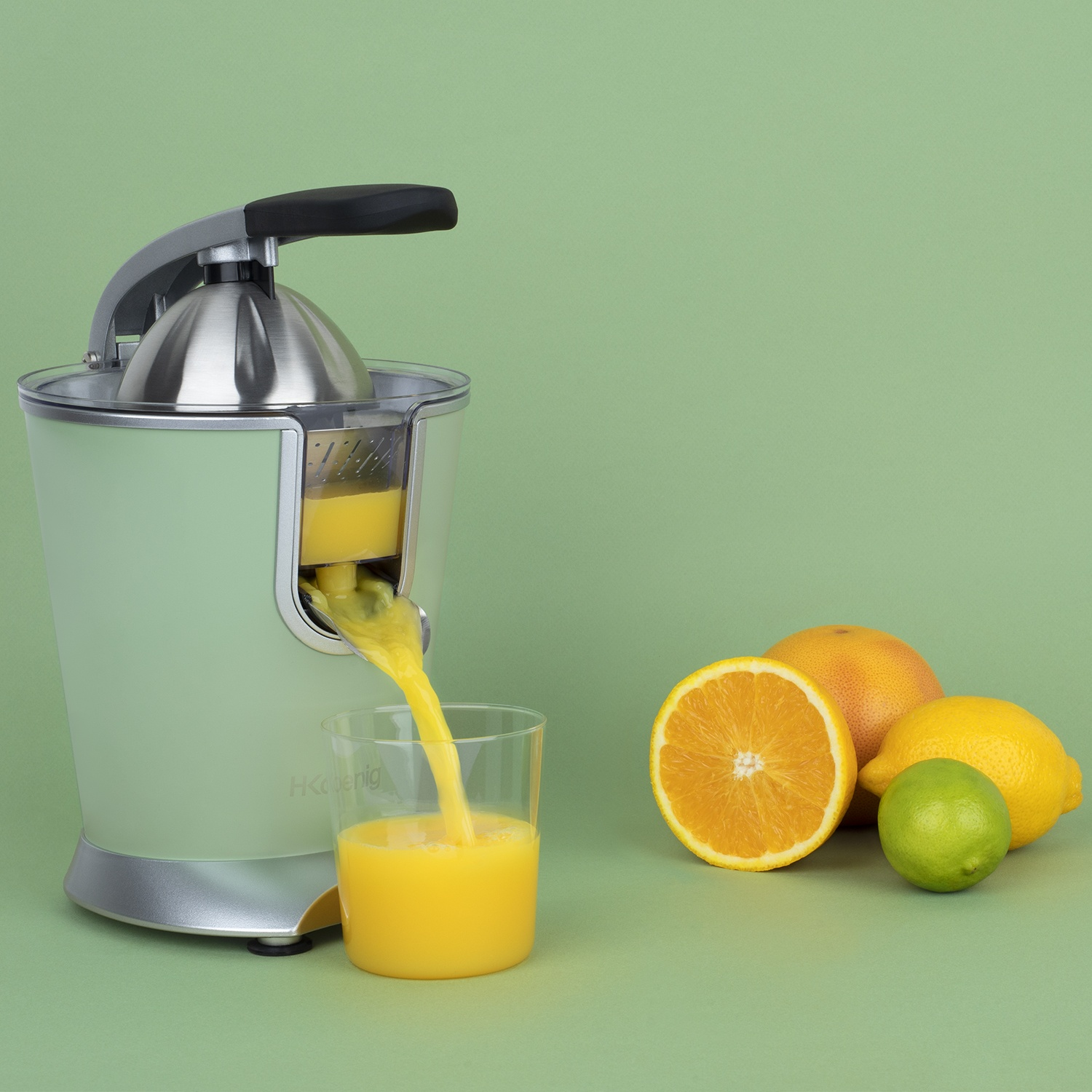 Succo versato da uno spremiagrumi in un bicchiere. Arance, limoni e lime accanto allo spremiagrumi su sfondo verde.
