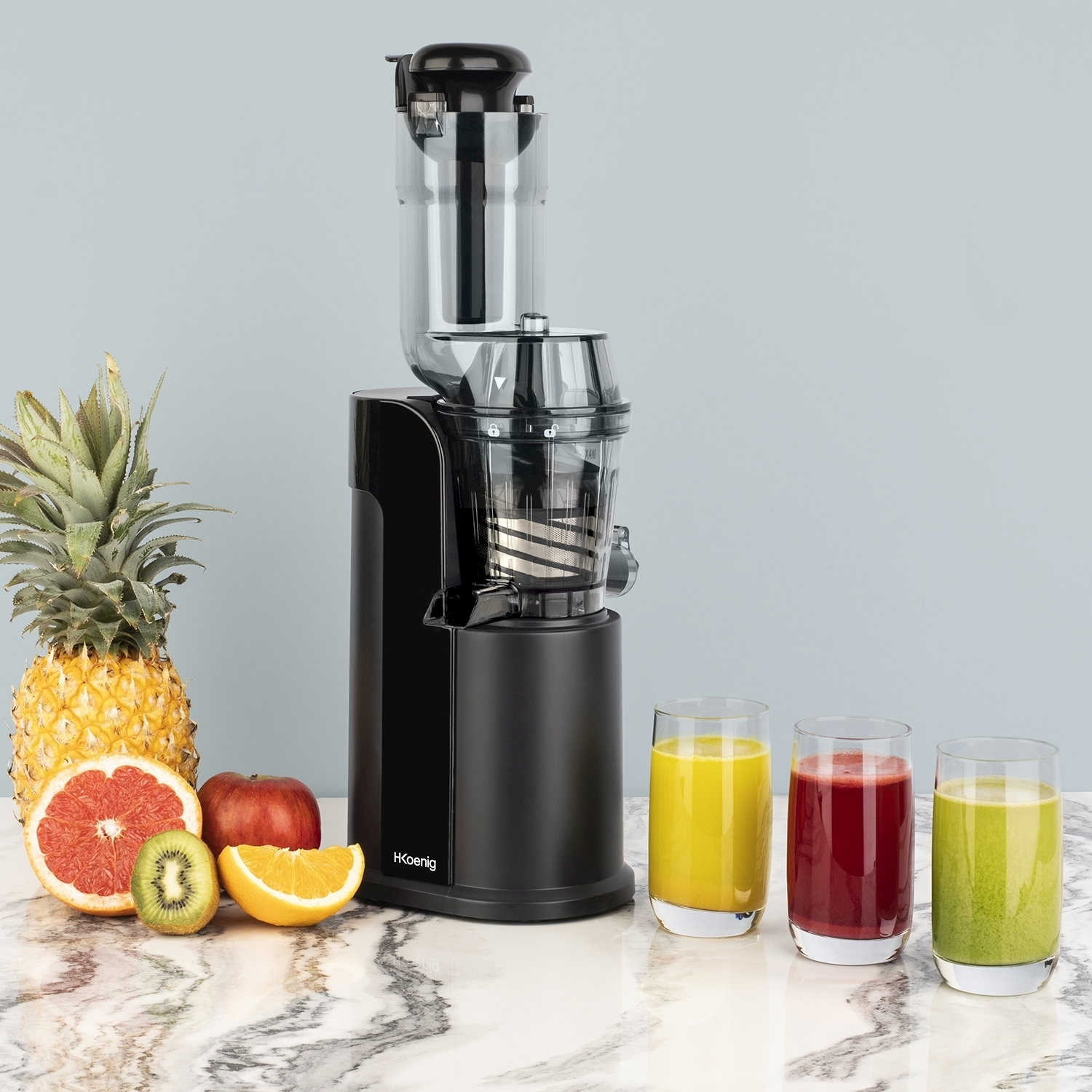 Un'estrattore di succo con frutta e tre bicchieri di succo su una superficie di marmo. La frutta comprende ananas, pompelmo, mela, arancia e kiwi.