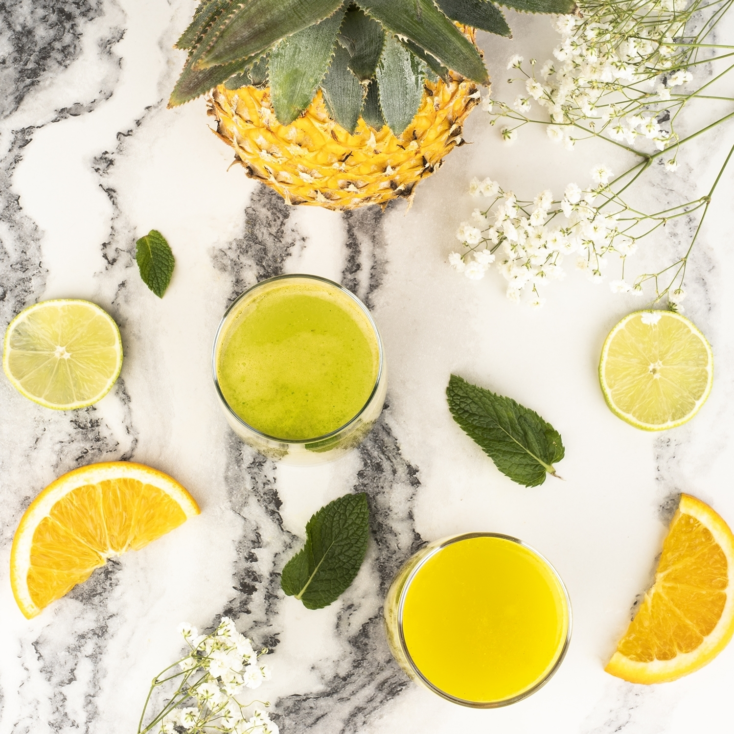 Vista dall'alto di frutta, bevande e fiori. Include ananas, lime, arancia, menta e due bicchieri di succo su marmo.