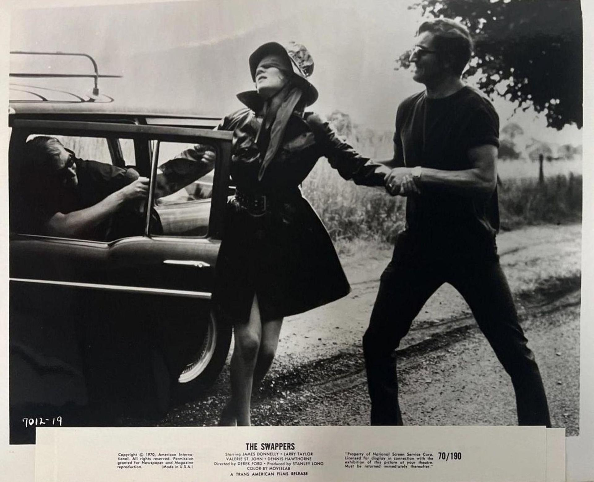 Schwarz-Weiß-Foto zeigt eine Frau mit Hut, die von einem Mann in der Nähe eines Autos geführt wird. Ein anderer Mann ist im Auto.