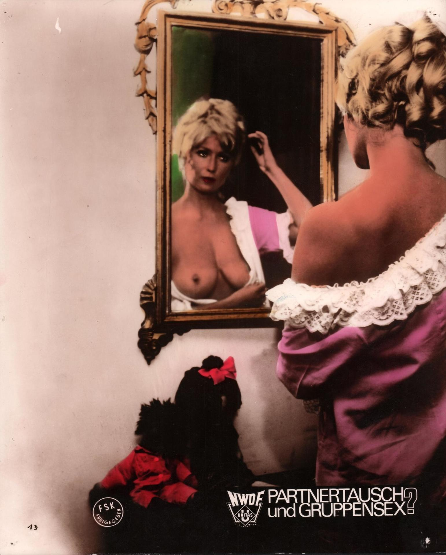 Frau im Spiegel, die sich die Haare richtet, trägt ein rosa Kleid. Im Spiegel ist ein nackter Oberkörper zu sehen. Schwarze Figur mit roter Schleife im Vordergrund.