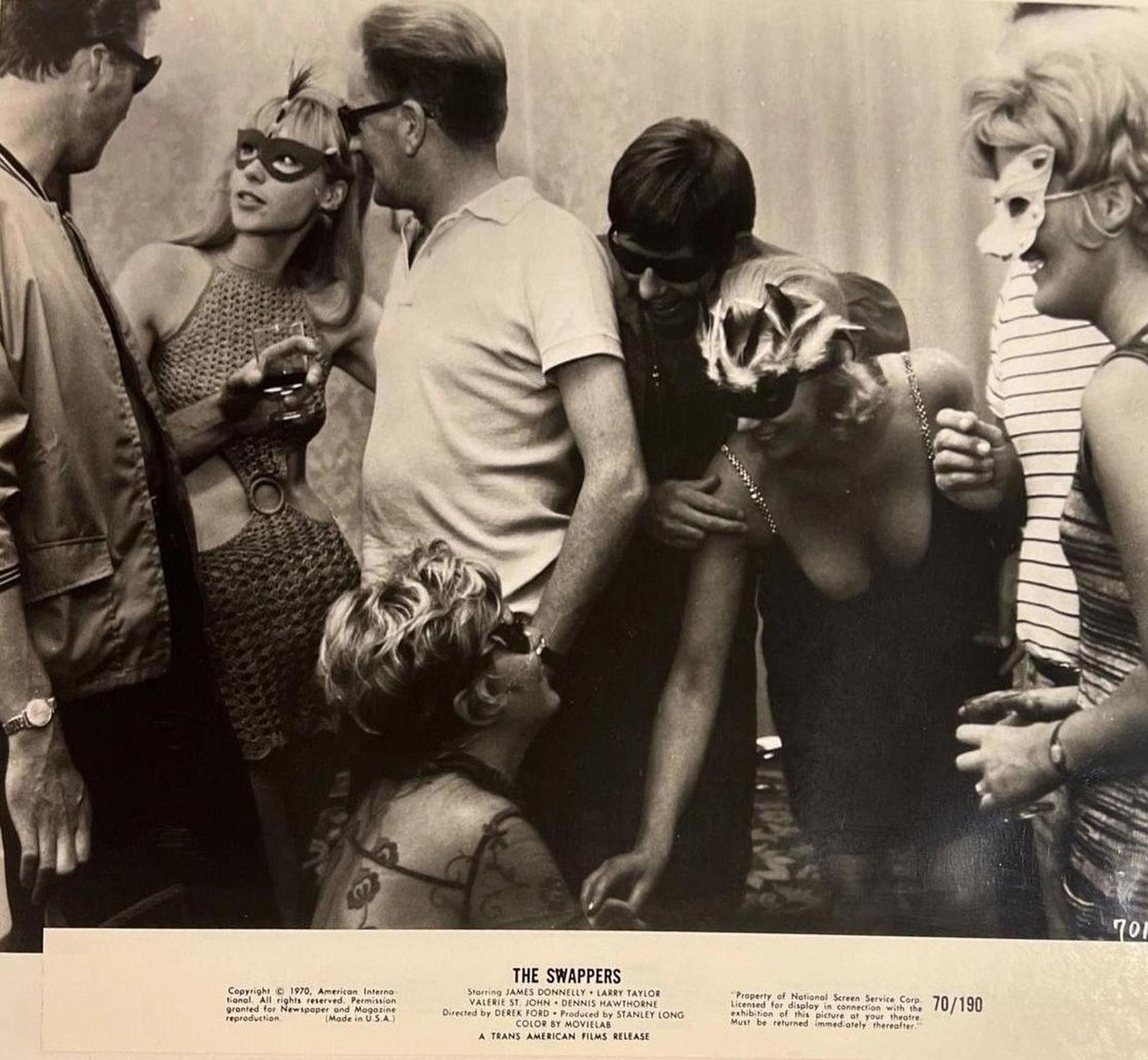 Gruppe von Menschen, einige mit Masken, bei einer Veranstaltung. Eine Frau in einem Neckholder-Kleid hält ein Getränk. Schwarz-Weiß-Foto.