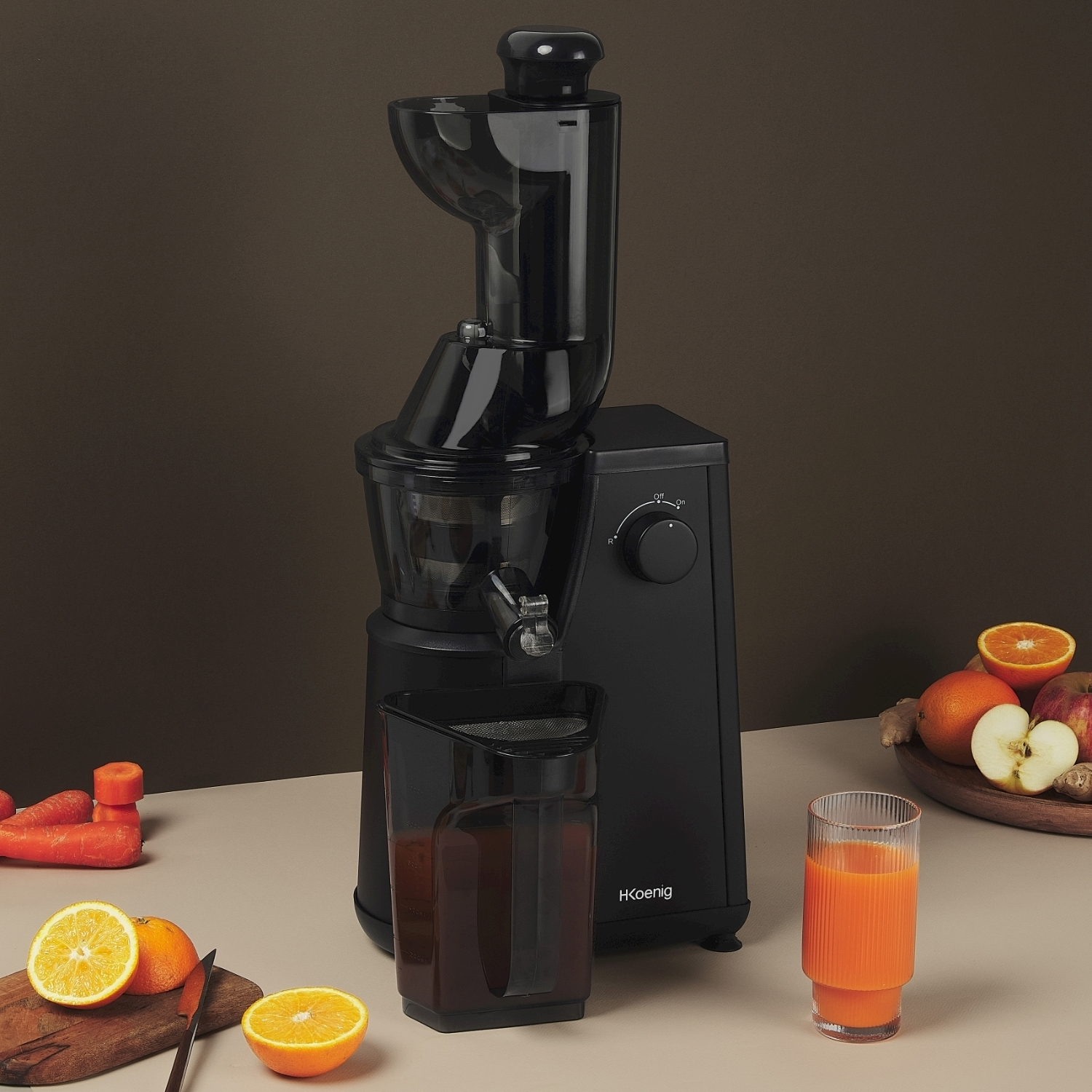 Centrifuga nera, bicchiere di succo, arance, carote, mele e zenzero su superficie beige, sfondo marrone.