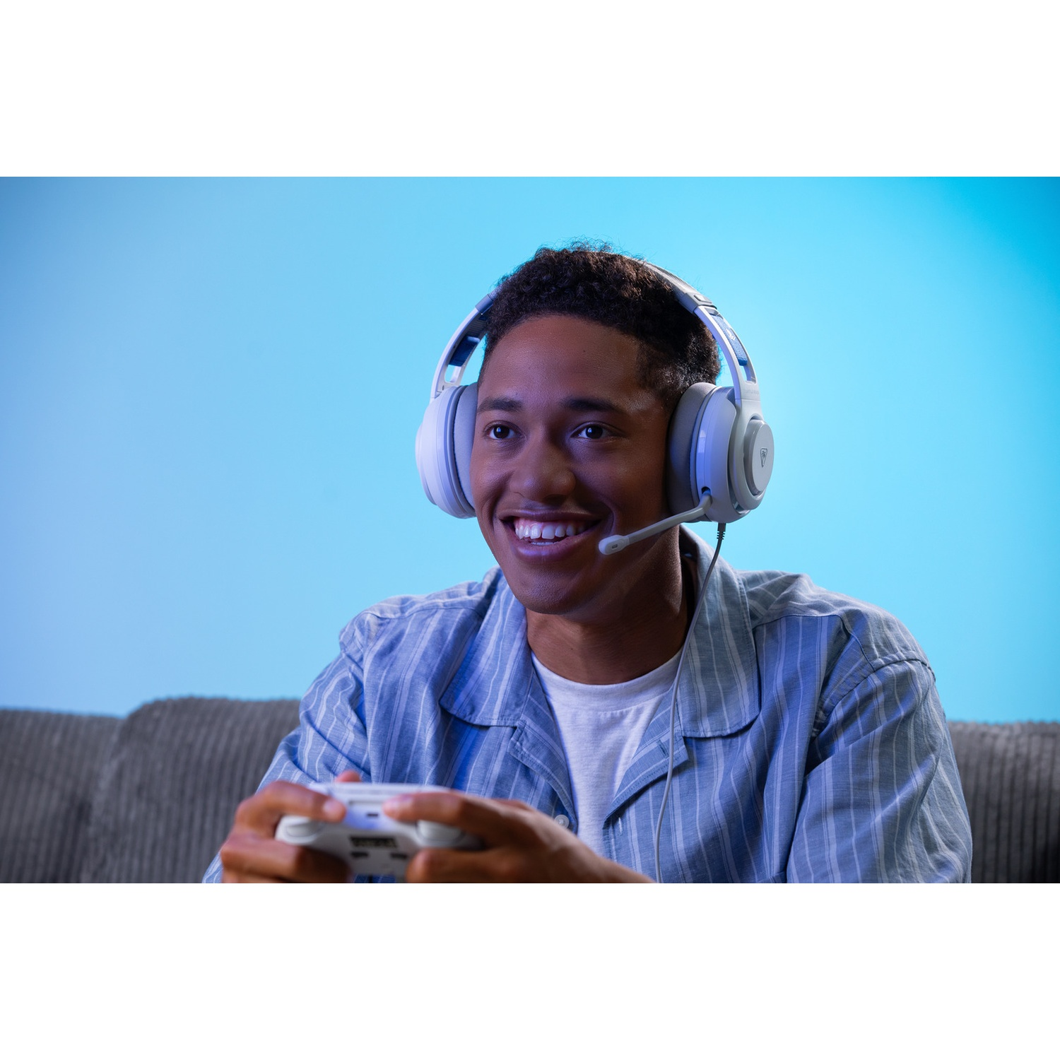 A young man with headphones is smiling while holding a game controller. He is sitting on a couch, with a blue background.