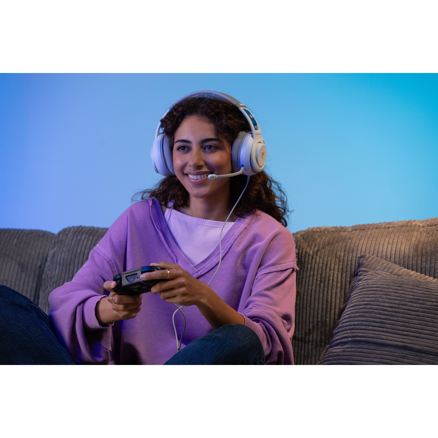 A woman wearing headphones and a headset smiles while holding a game controller, sitting on a couch.