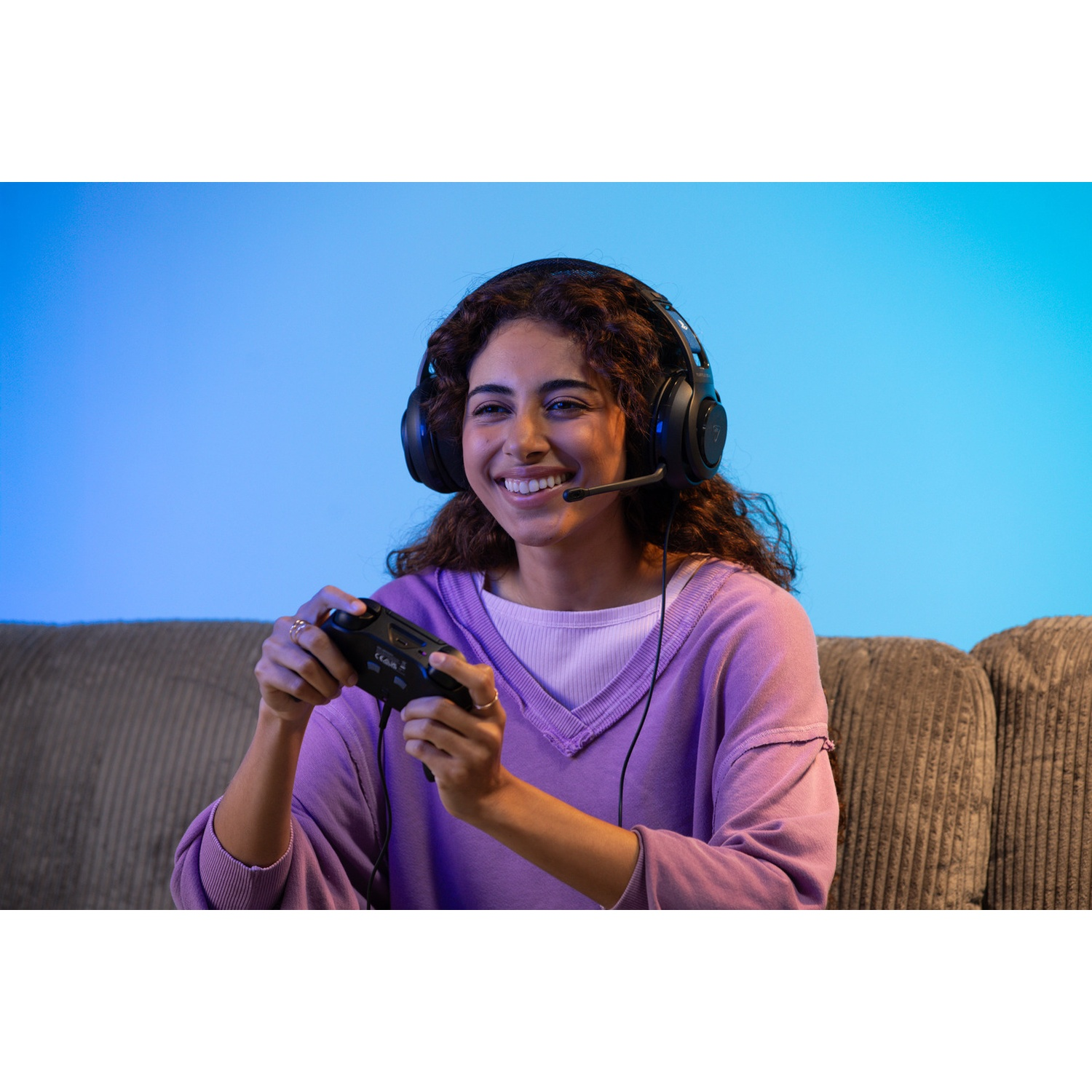 Een vrouw glimlacht tijdens het gamen. Ze draagt een headset en houdt een controller vast. Een paarse trui en een blauwe achtergrond zijn zichtbaar.