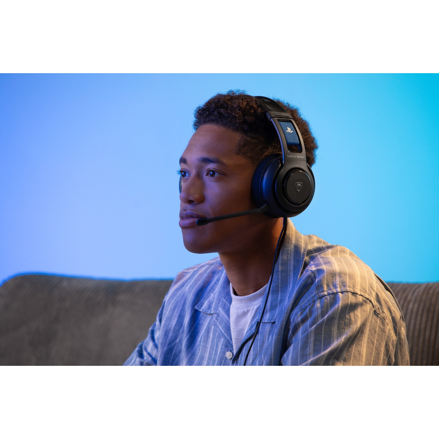 Een jonge man met een headset en microfoon zit op een bank. Hij kijkt naar links, met een blauwe achtergrond.