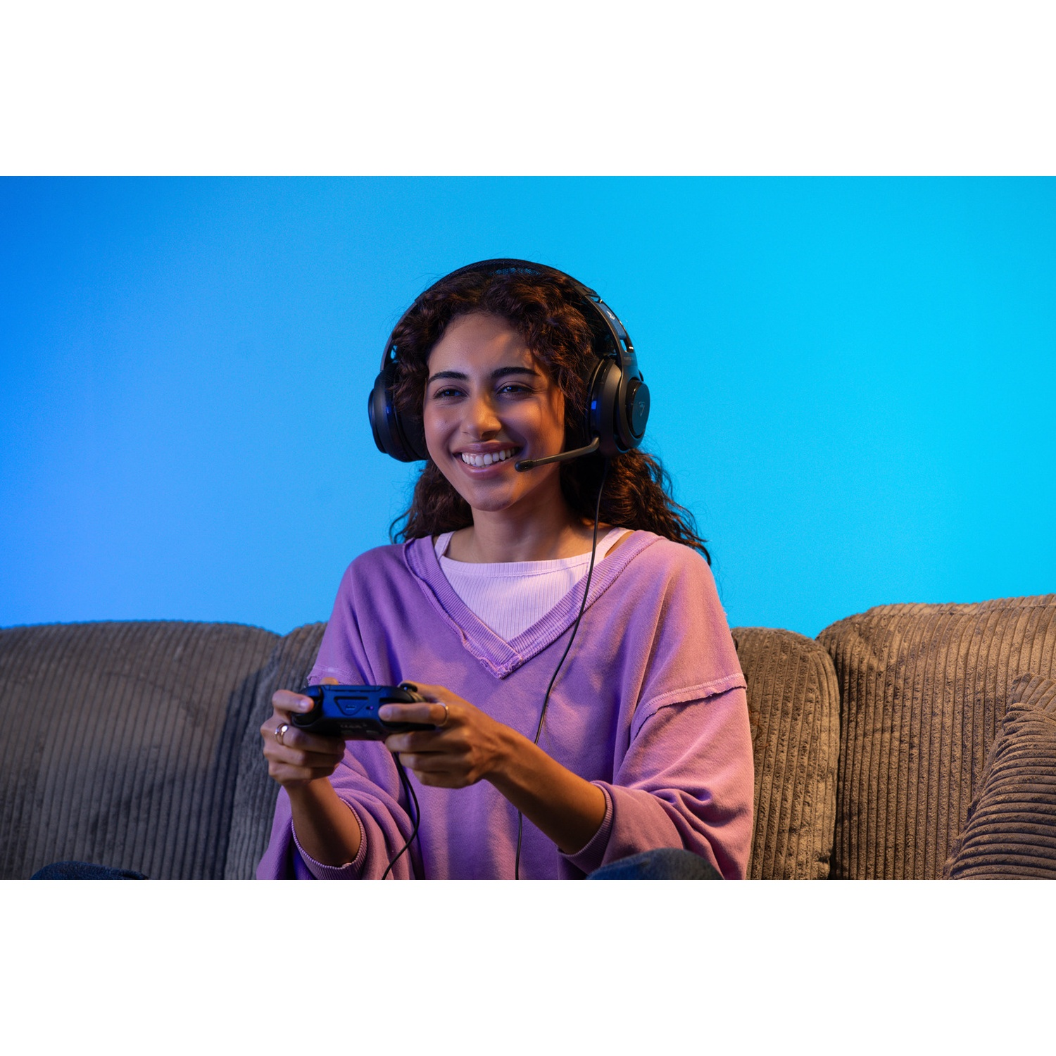 Een vrouw met een headset glimlacht terwijl ze een spel speelt. Ze houdt een controller vast en zit op een bank met een blauwe achtergrond.