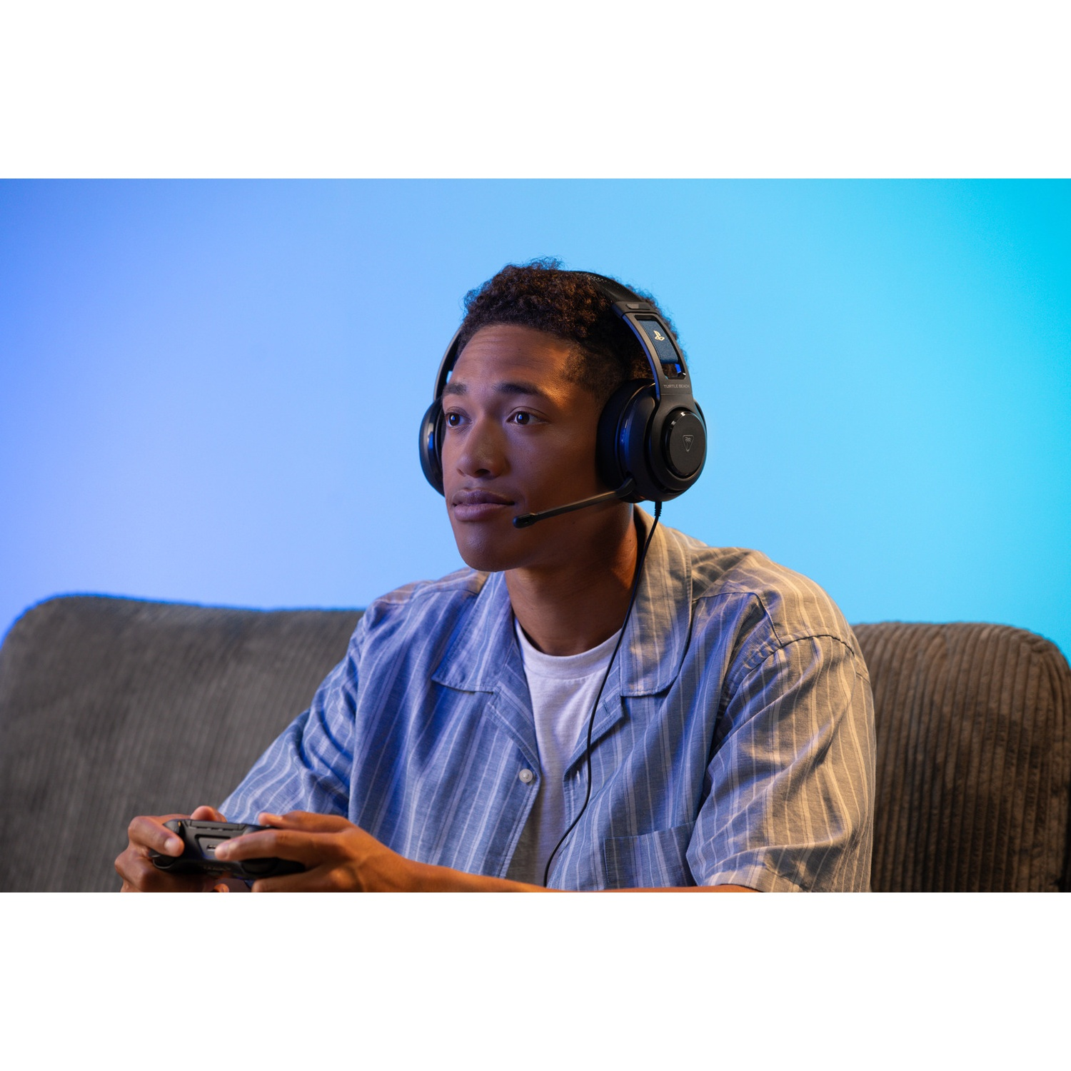 Een jonge man met een headset speelt een videogame terwijl hij een controller vasthoudt. Hij zit op een bank. Blauwe achtergrond.