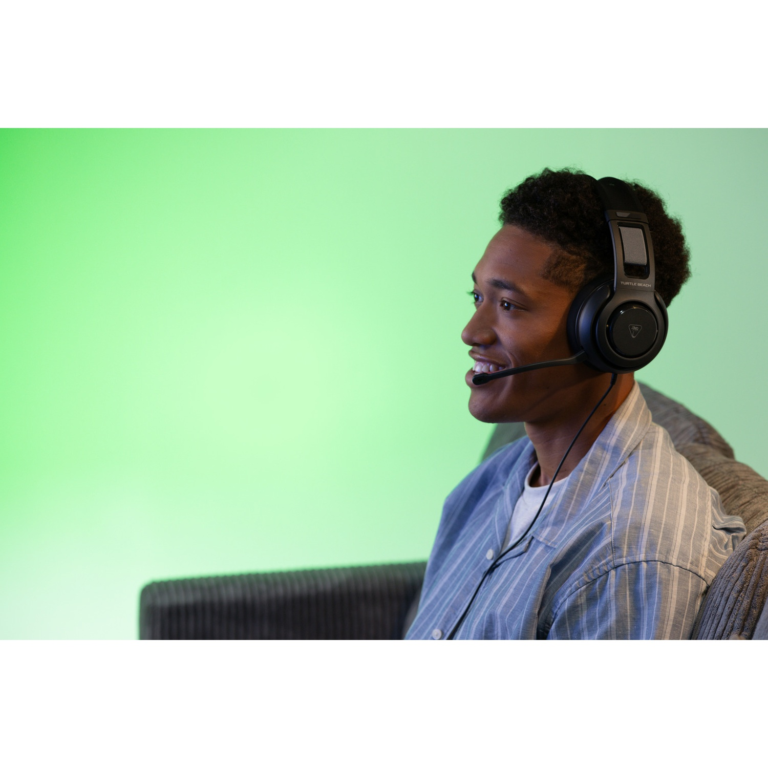 A young man wearing a gaming headset smiles. He sits on a sofa, with a green background.