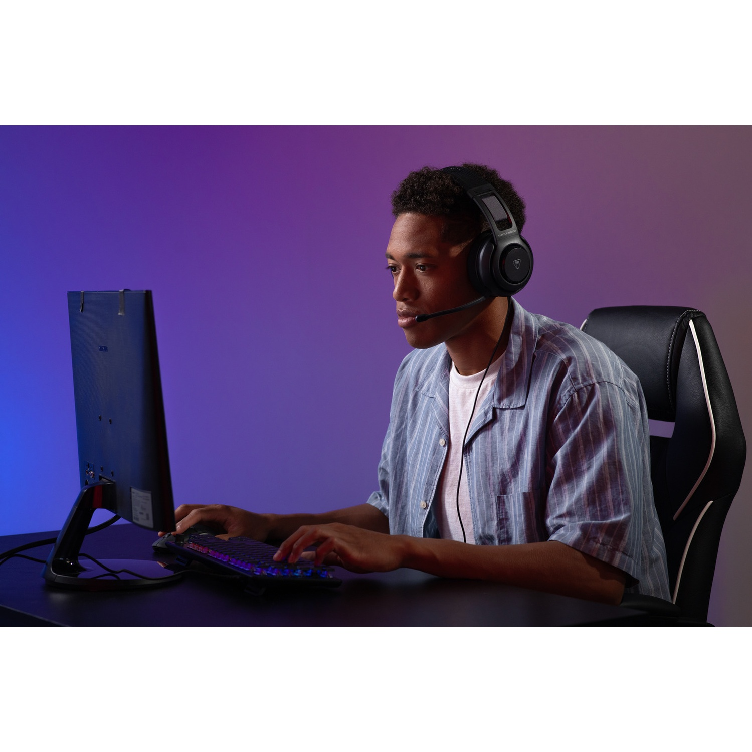 A man wearing a headset is gaming at a desk. He's in front of a monitor and keyboard, sitting in a black chair.