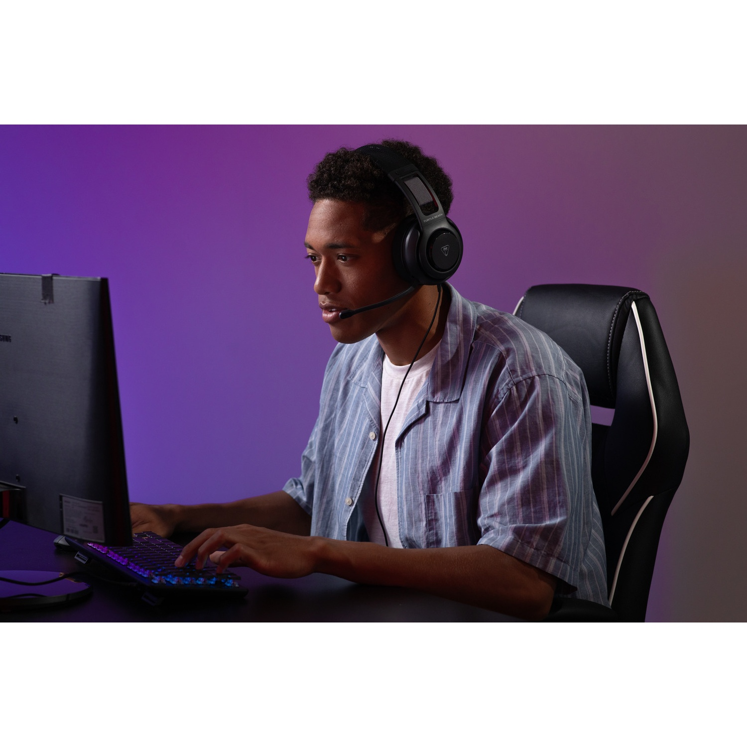 A young man wearing a headset is gaming at a desk. He's in a gaming chair, looking at the screen. Keyboard is lit up.