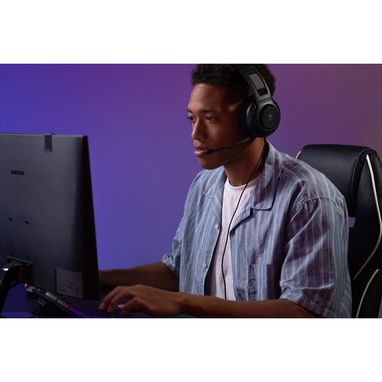 A young man wearing a headset sits at a computer, typing on the keyboard. A monitor is behind him and he's in a chair.